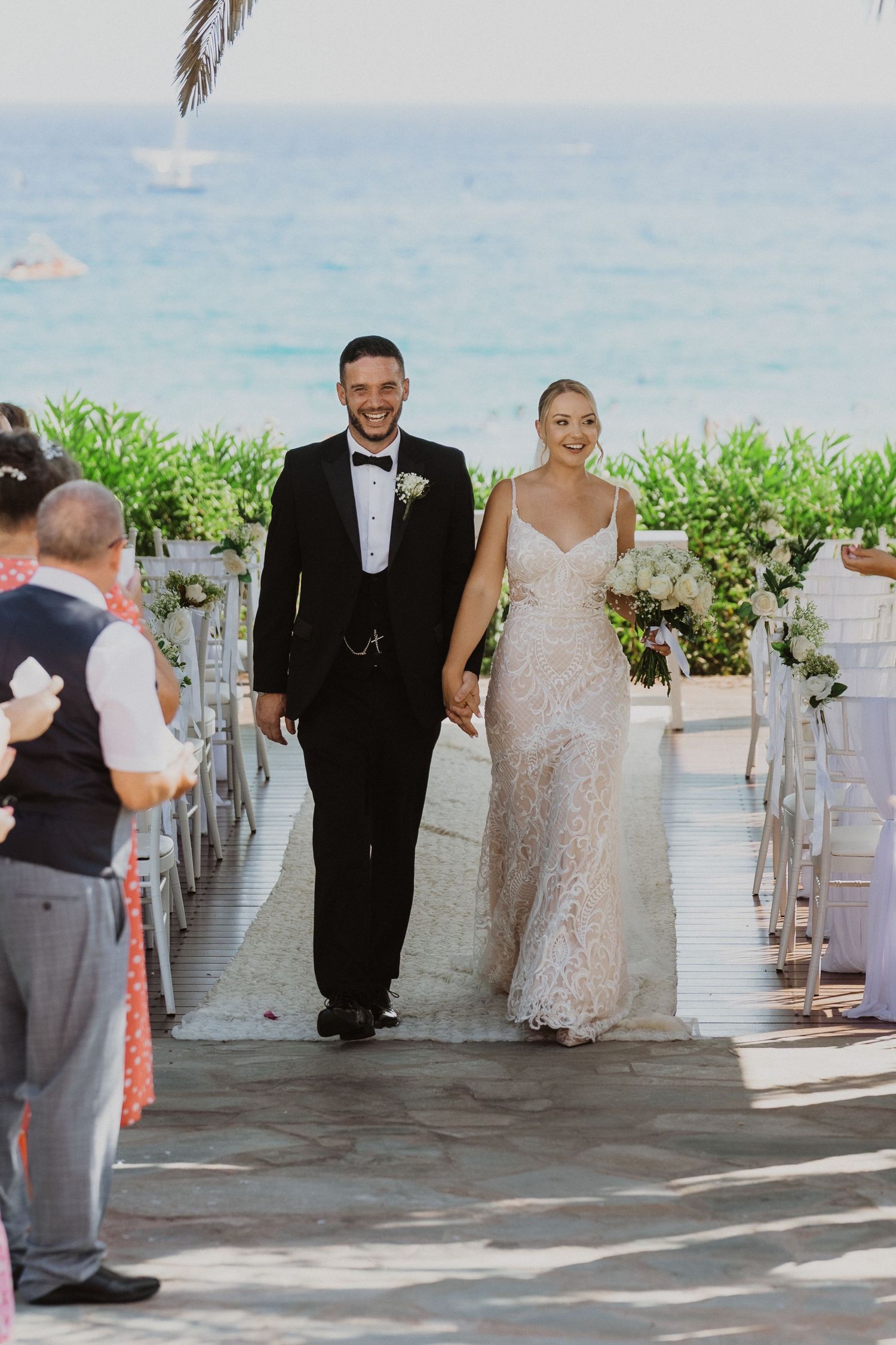 Wedding couple walks along an oceanfront terrace with palm trees and white decor for their ceremony.