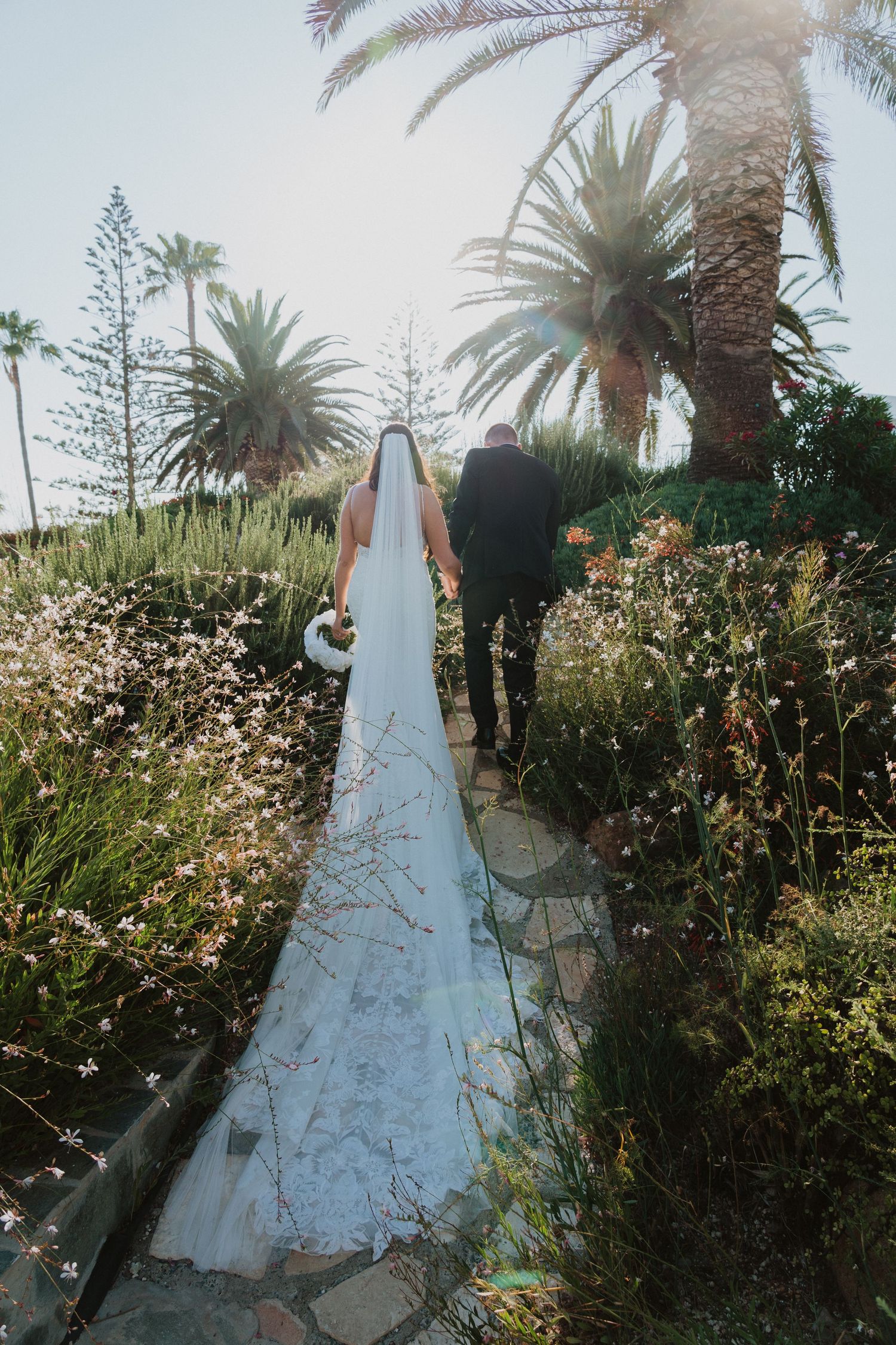 A couple walks down a garden path with palm trees and blooming flowers on a sunny day.