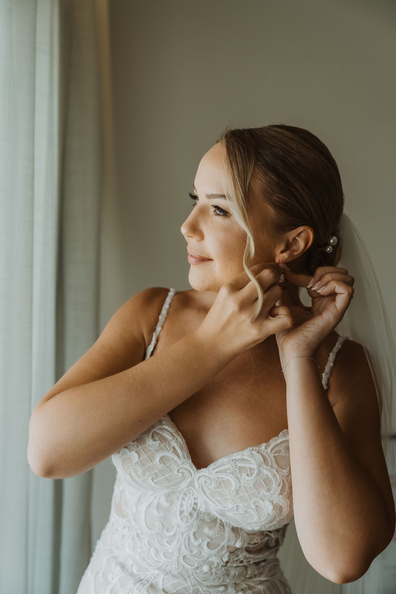 Bride puts on earrings while getting ready in a white lace wedding dress by a window.