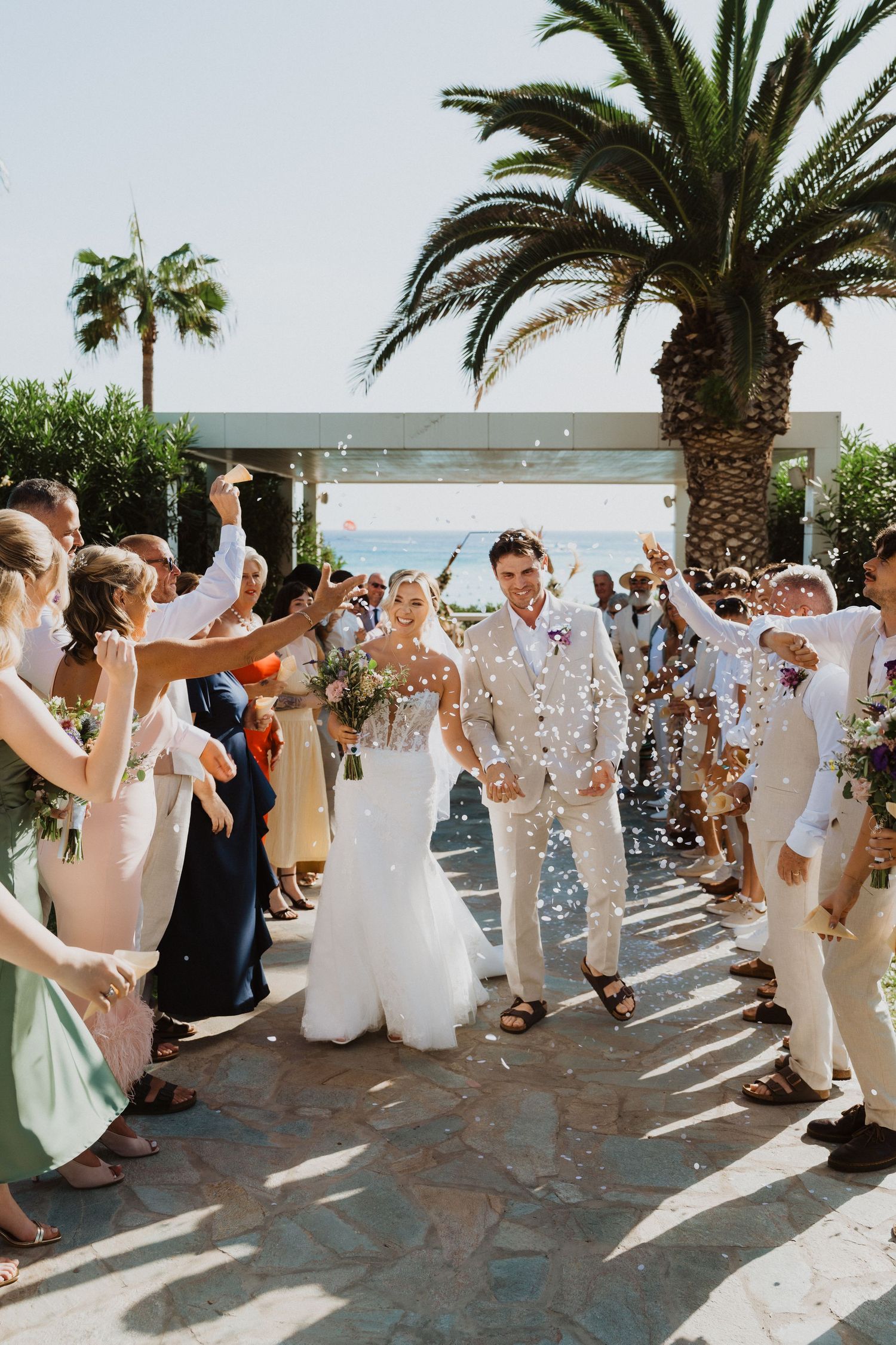 Wedding party exits under a shower of confetti along a palm-lined walkway at a beach venue.