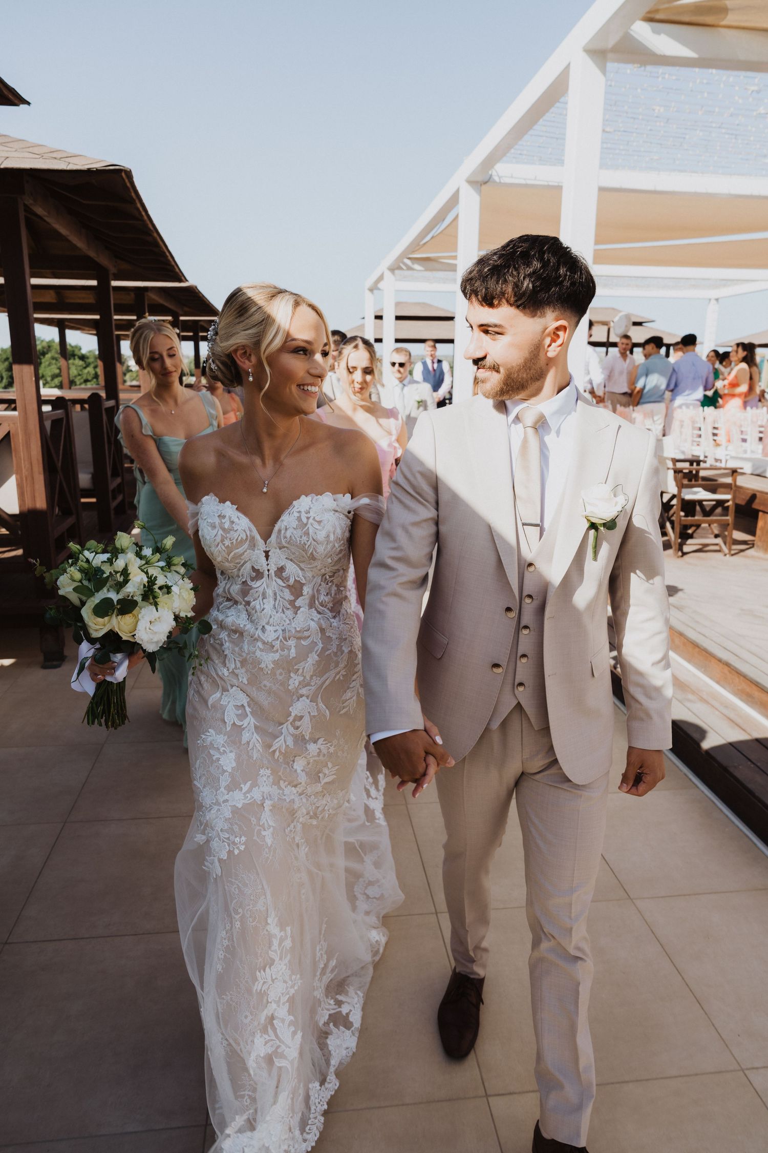 Newlyweds walking hand in hand on their wedding day at a seaside venue.