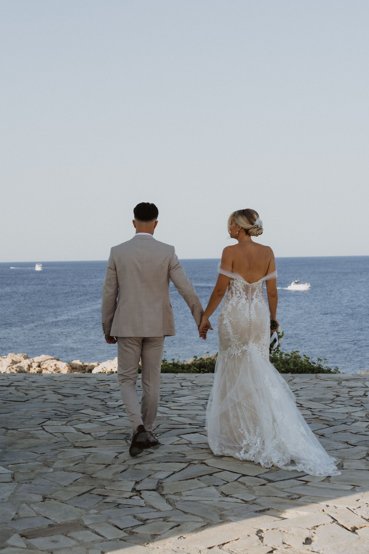 Wedding couple strolls on stone terrace with ocean view in background.
