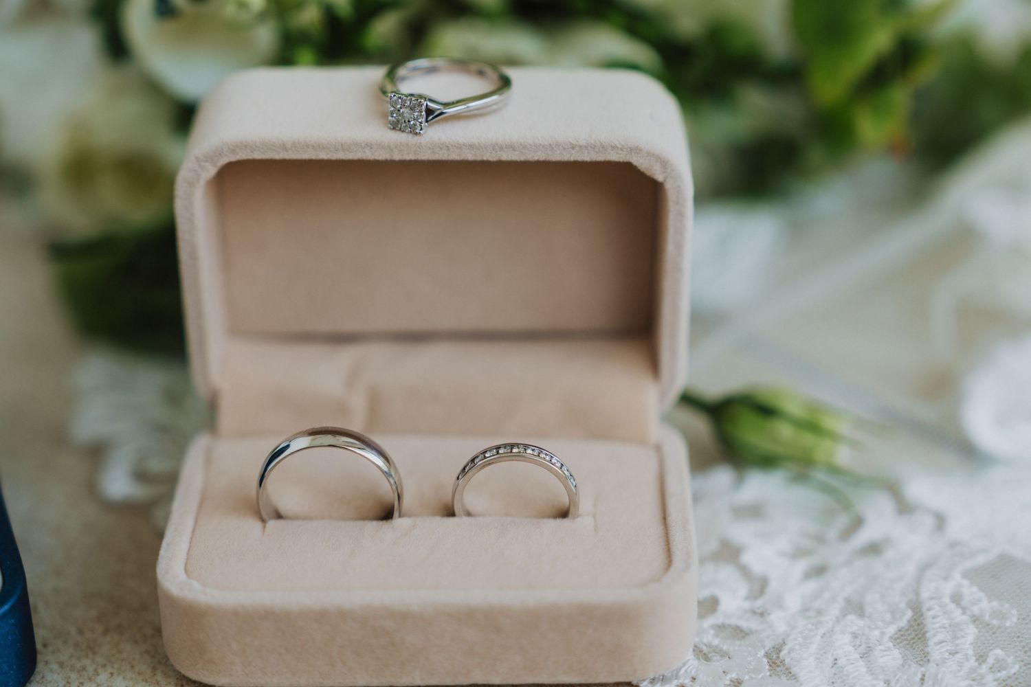 Wedding rings nestled in a beige velvet ring box surrounded by white flowers.