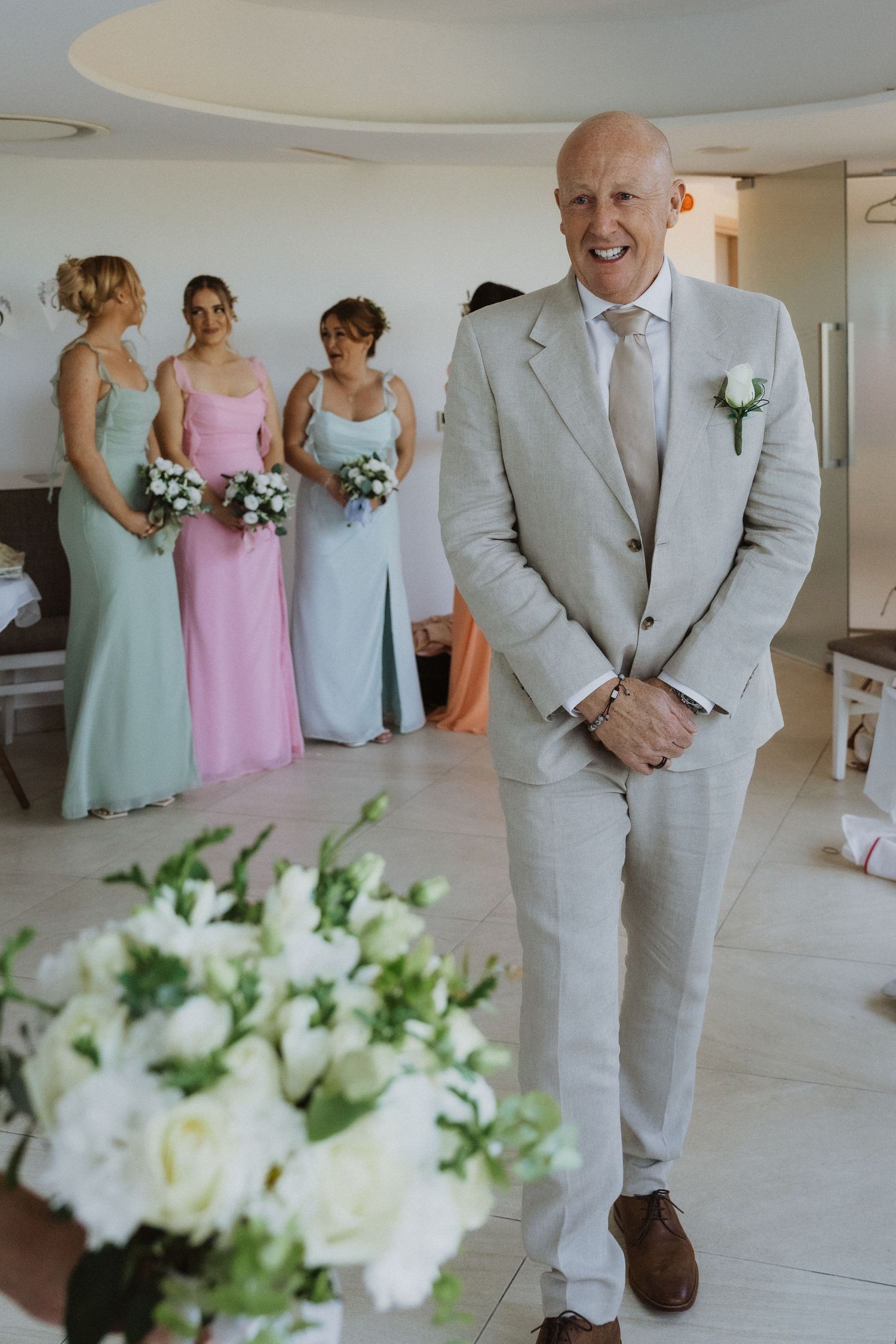 Wedding party members in pastel dresses and grey suits gather in a bright indoor space with white floral arrangements.