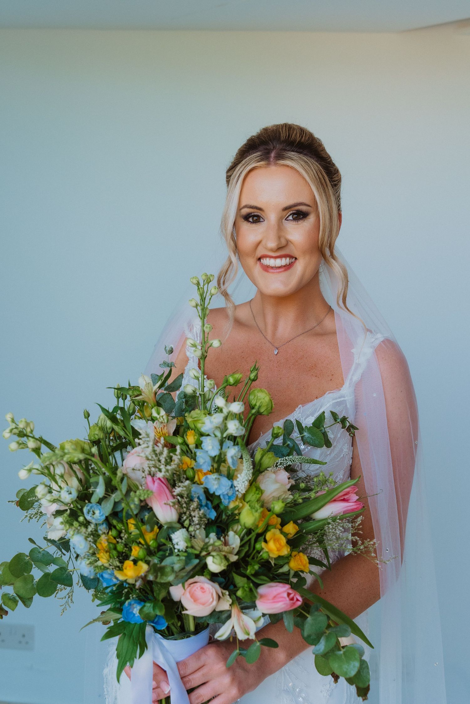 Beautiful wedding bouquet featuring pink, yellow and white flowers with lush greenery against a light blue background.