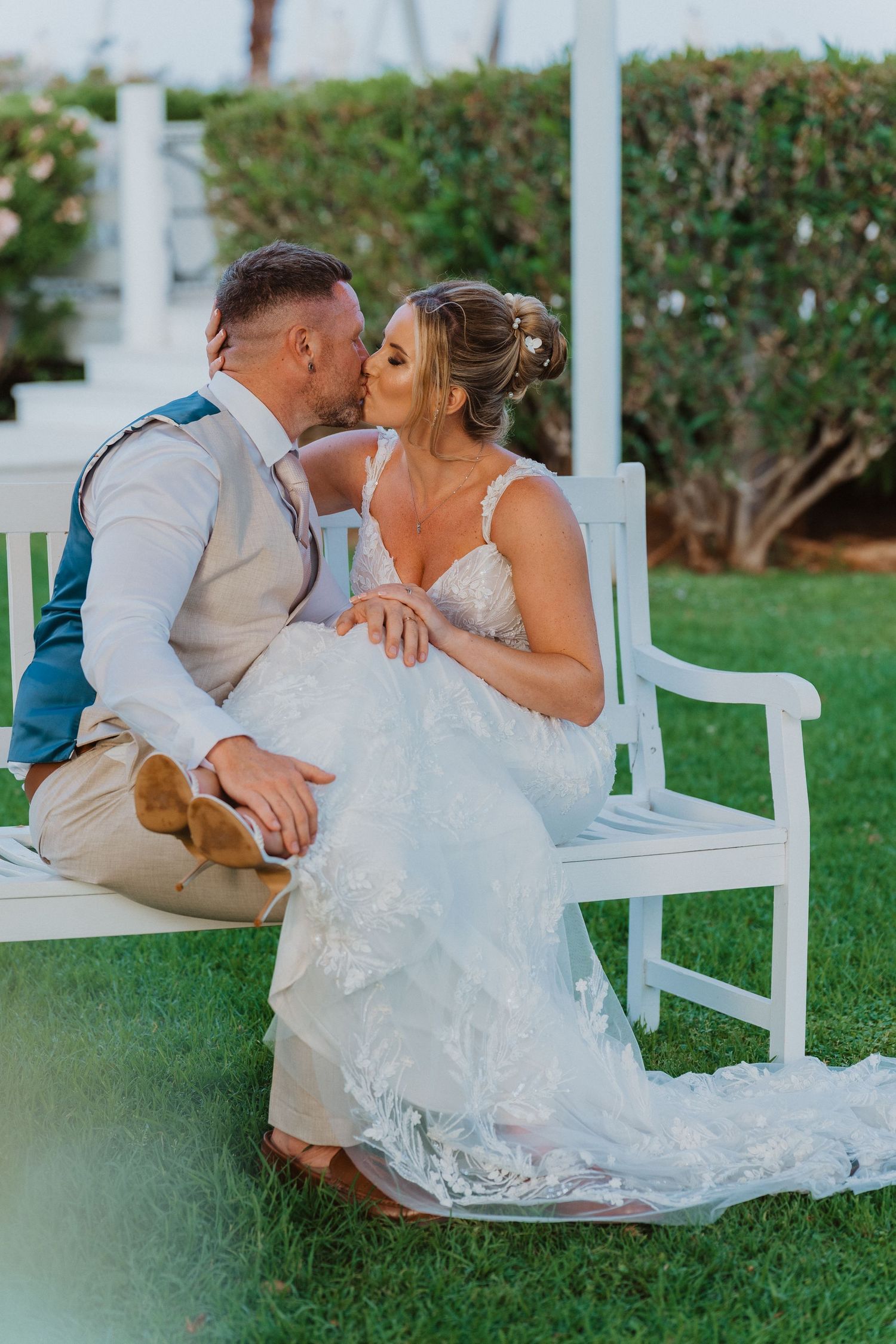 A couple in wedding attire shares romantic moments while sitting together on a white wooden bench in a garden setting.