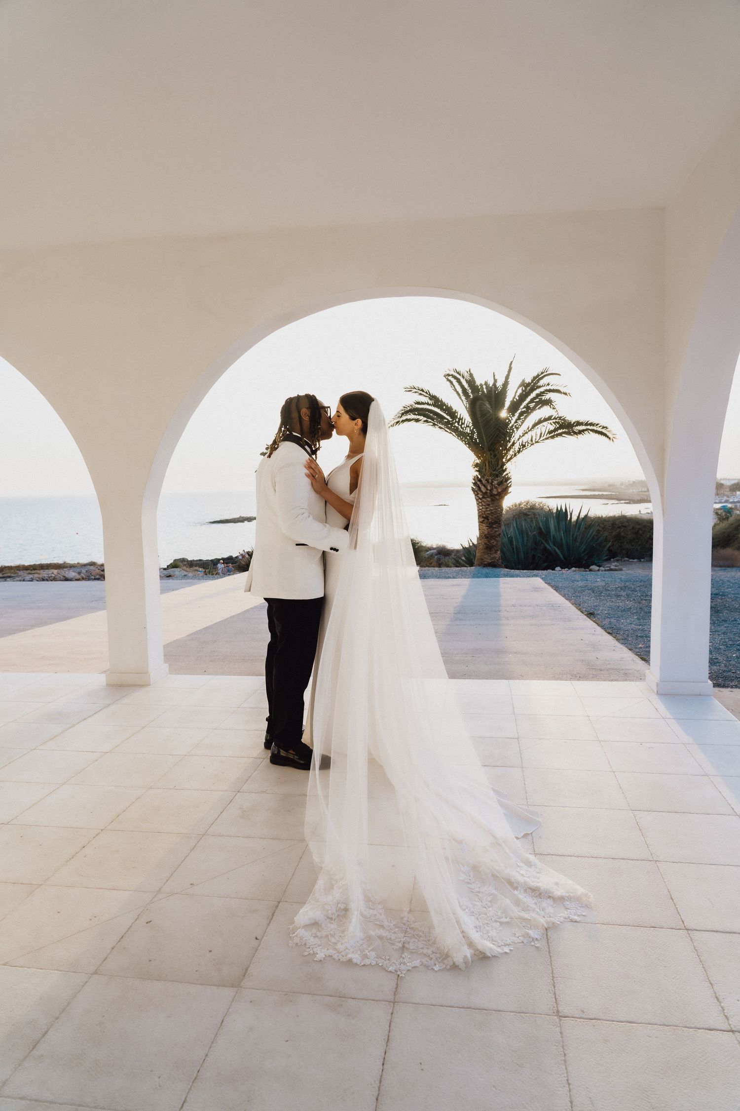 Intimate wedding moment under white arched hallway with palm tree and sea view.
