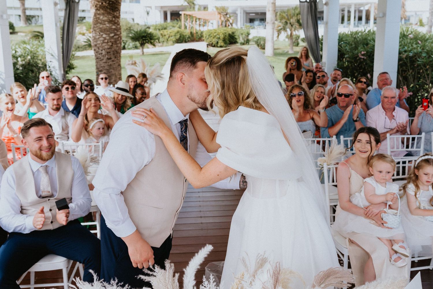 Newlyweds share first kiss during beachside ceremony as guests cheer.