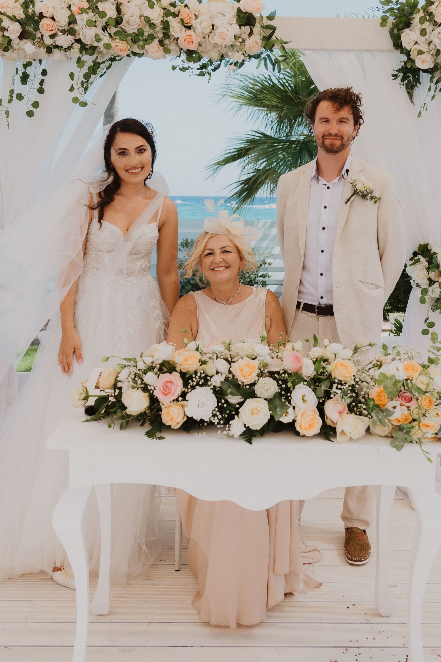 Three people pose at elegant beach wedding with floral arch and ocean backdrop.