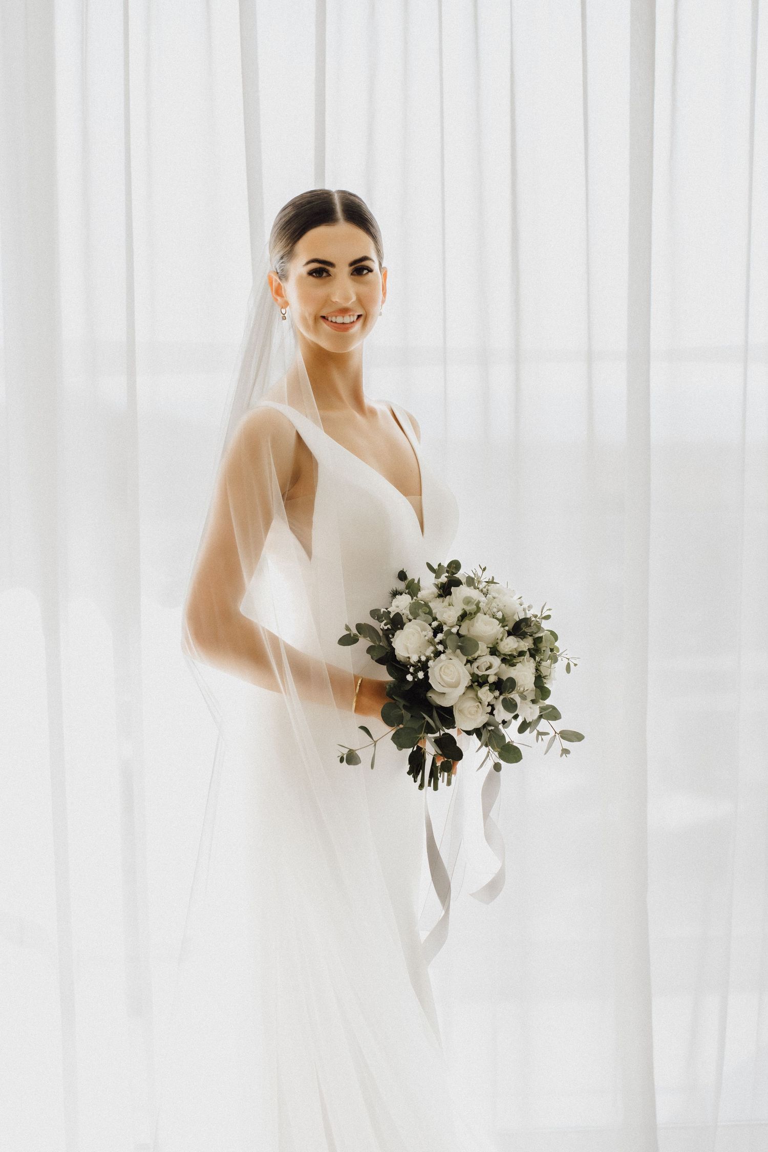 Bride in sleek white V-neck wedding dress poses with white and green bouquet against sheer curtains.