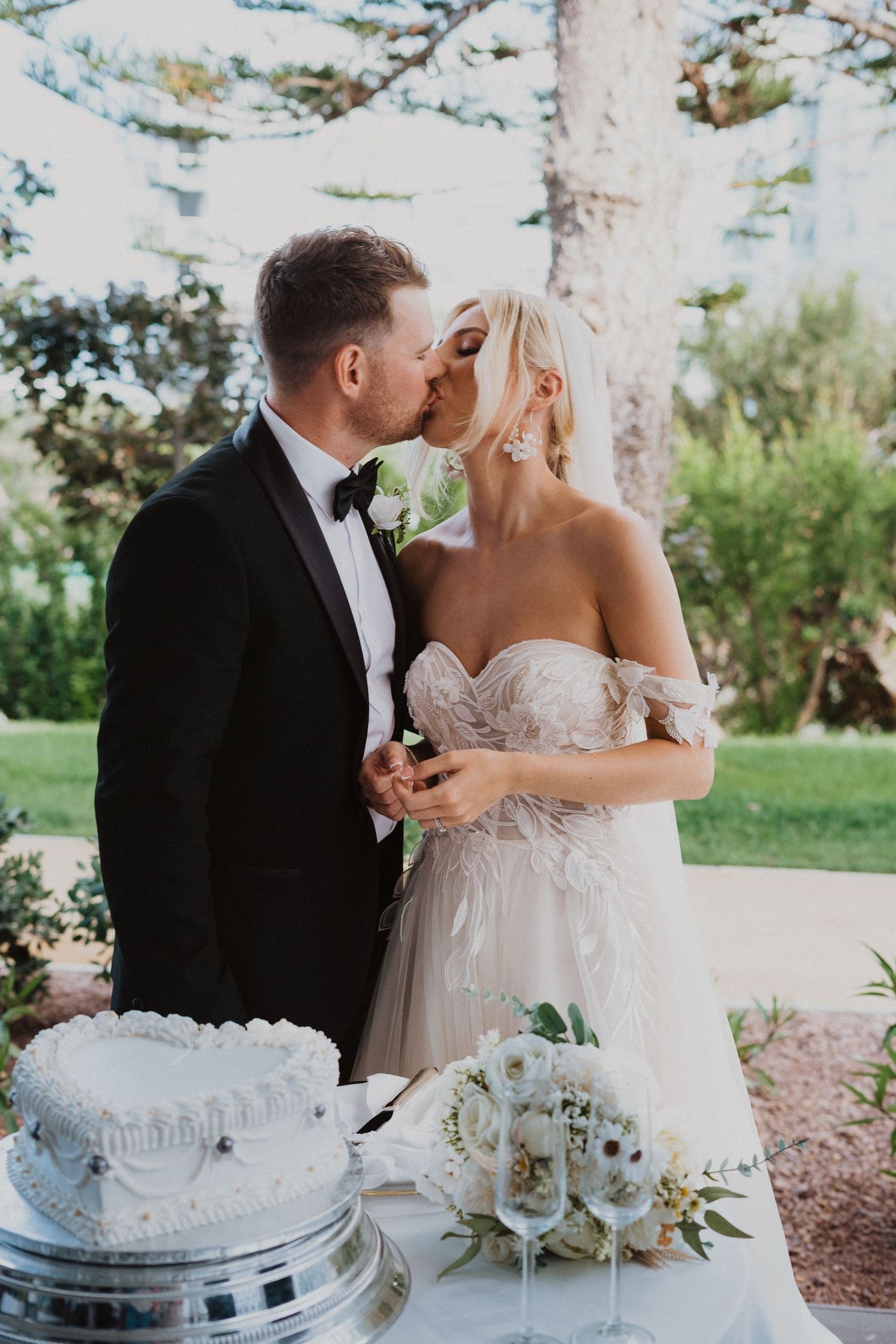 A romantic wedding moment with newlyweds sharing a kiss next to an elegant white wedding cake and floral arrangements.