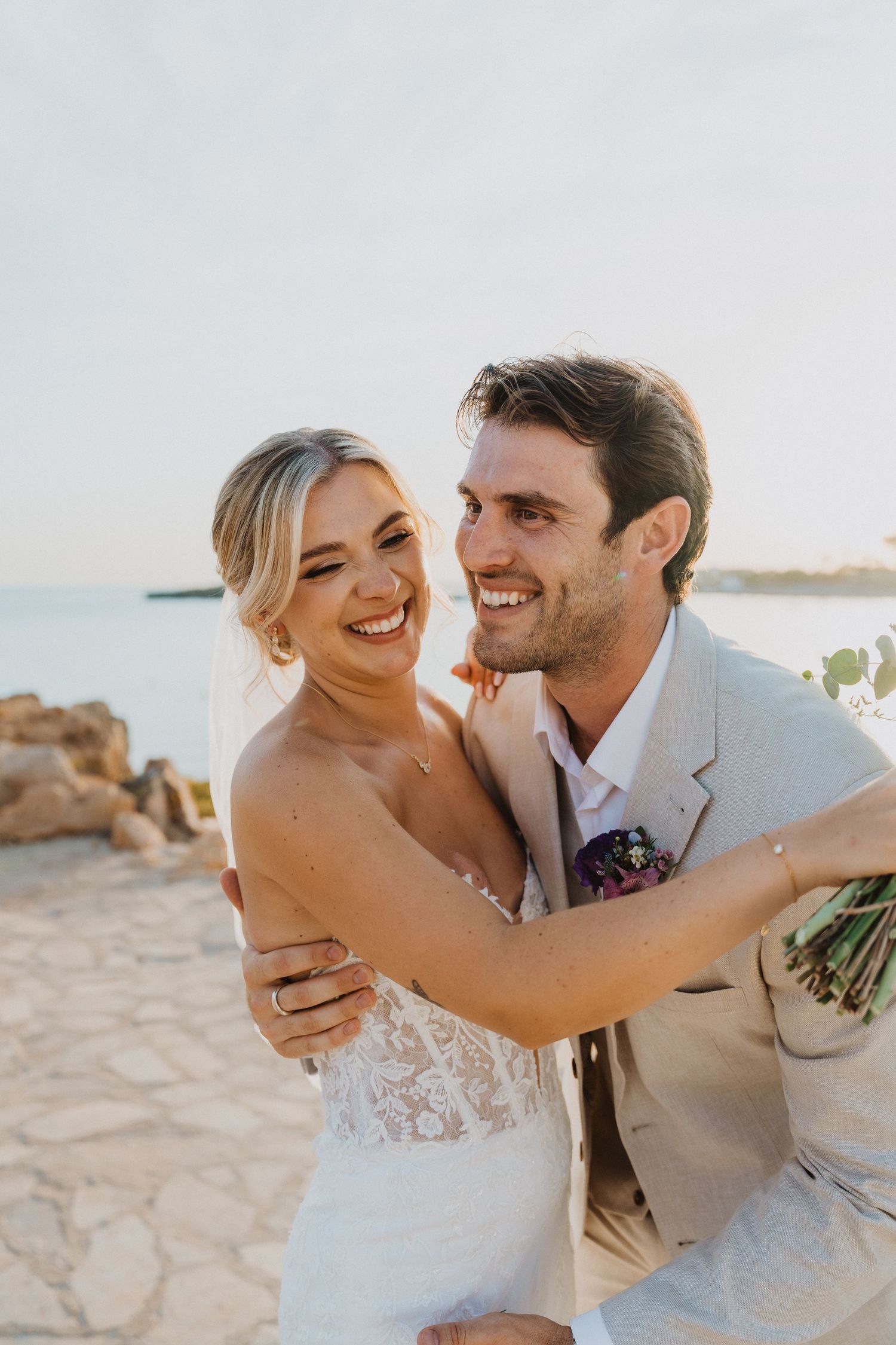 Playful wedding portraits show couple laughing and embracing on rocky shoreline.