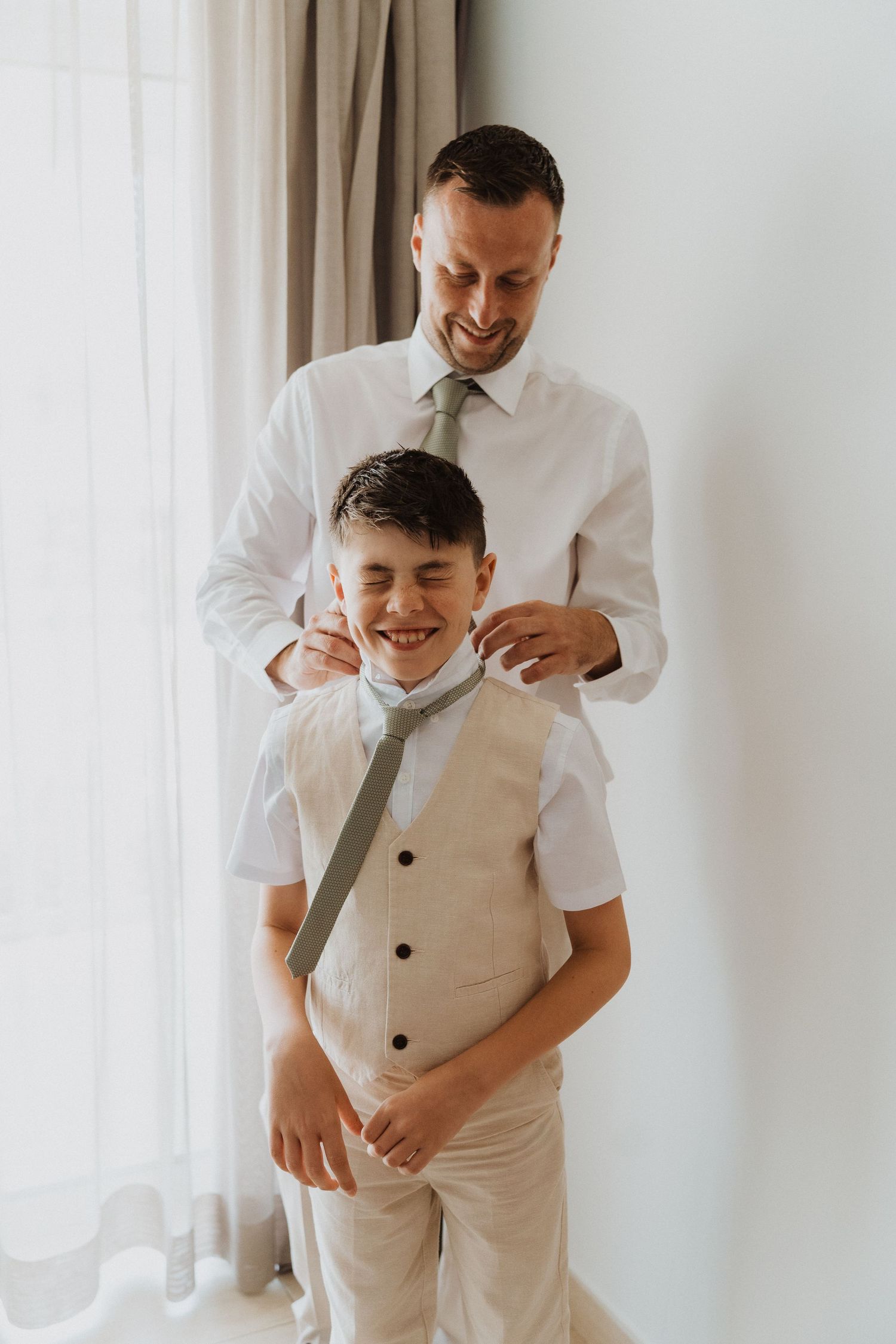 A sweet father and son moment as they both wear coordinating formal attire in white and beige against a light background.