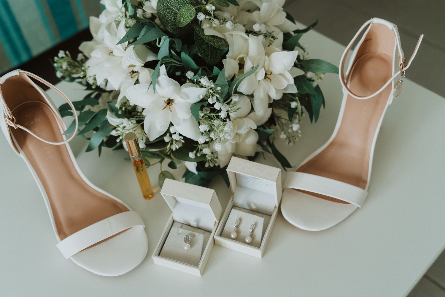 White bridal heels next to ring boxes and an elegant white floral bouquet with greenery arranged on table.