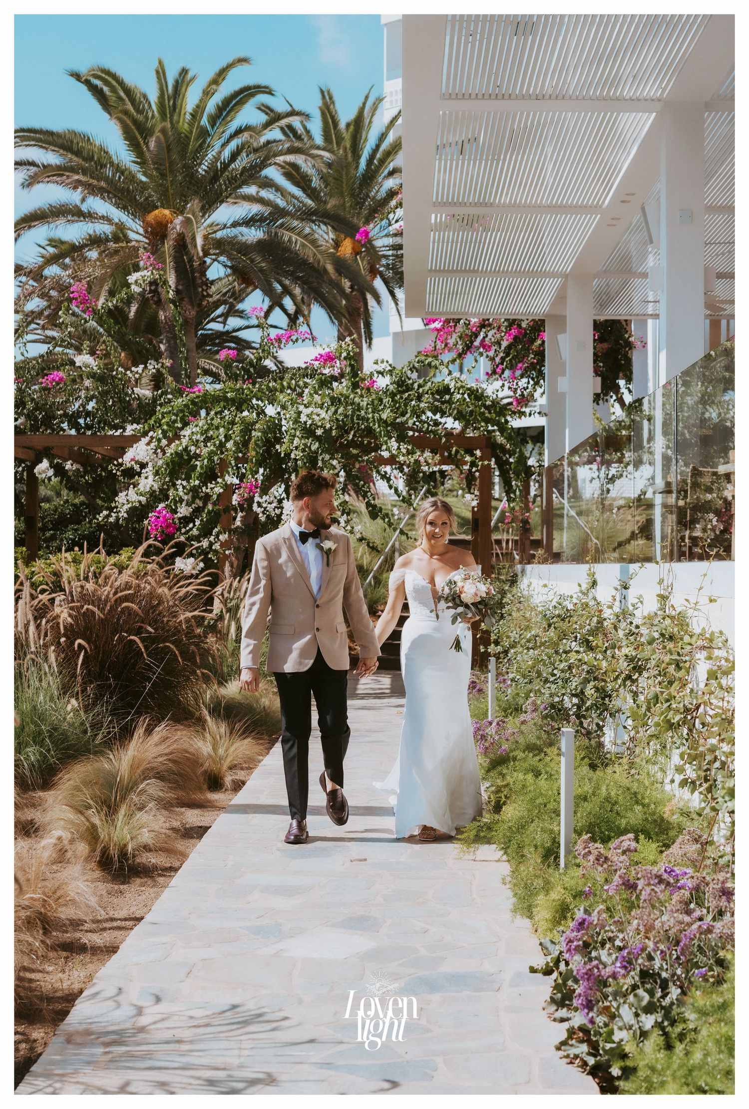 Newlyweds walk along a garden path lined with palm trees and colorful flowers.