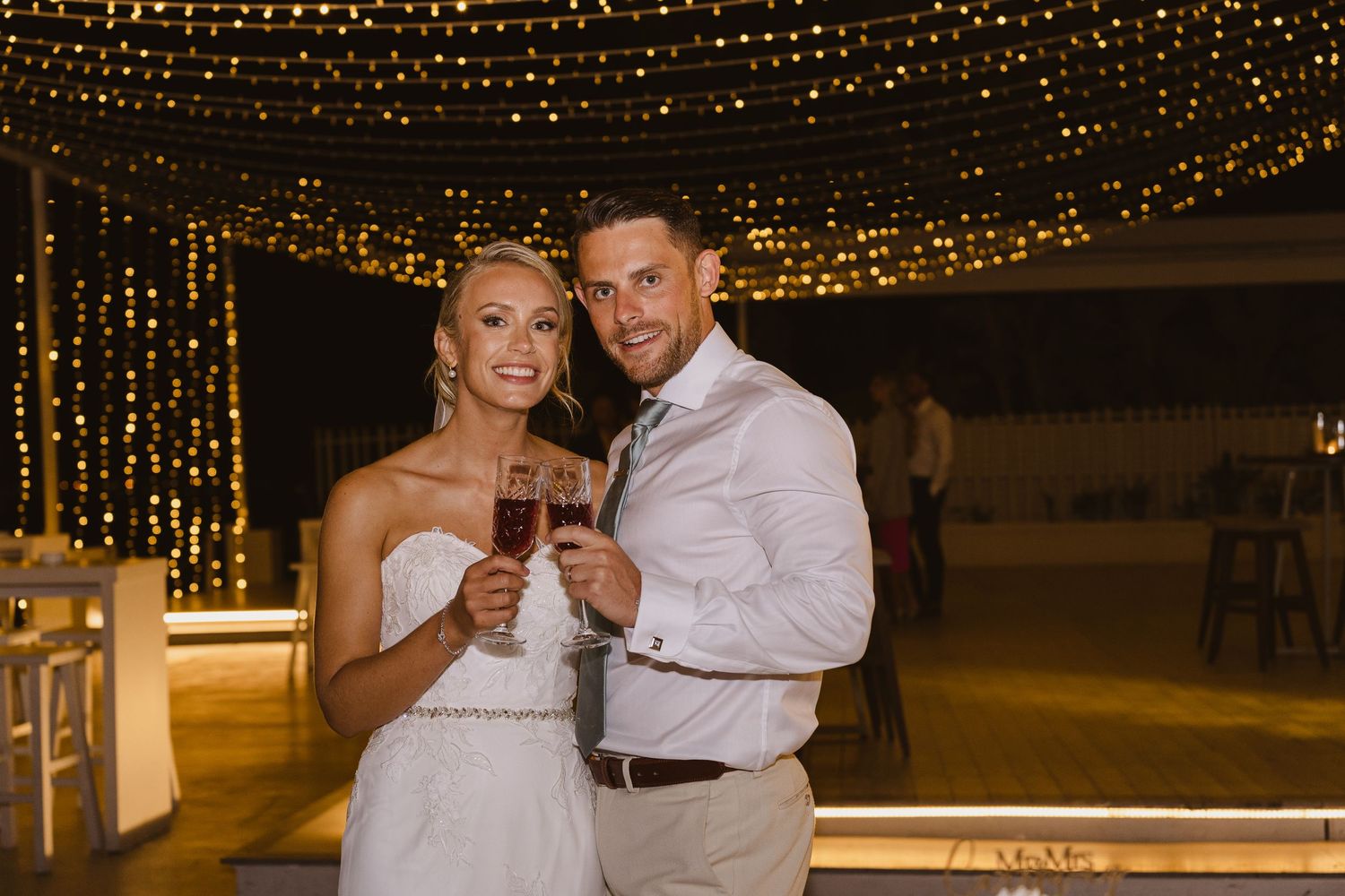 Wedding couple holds champagne glasses while celebrating under a magical canopy of string lights.