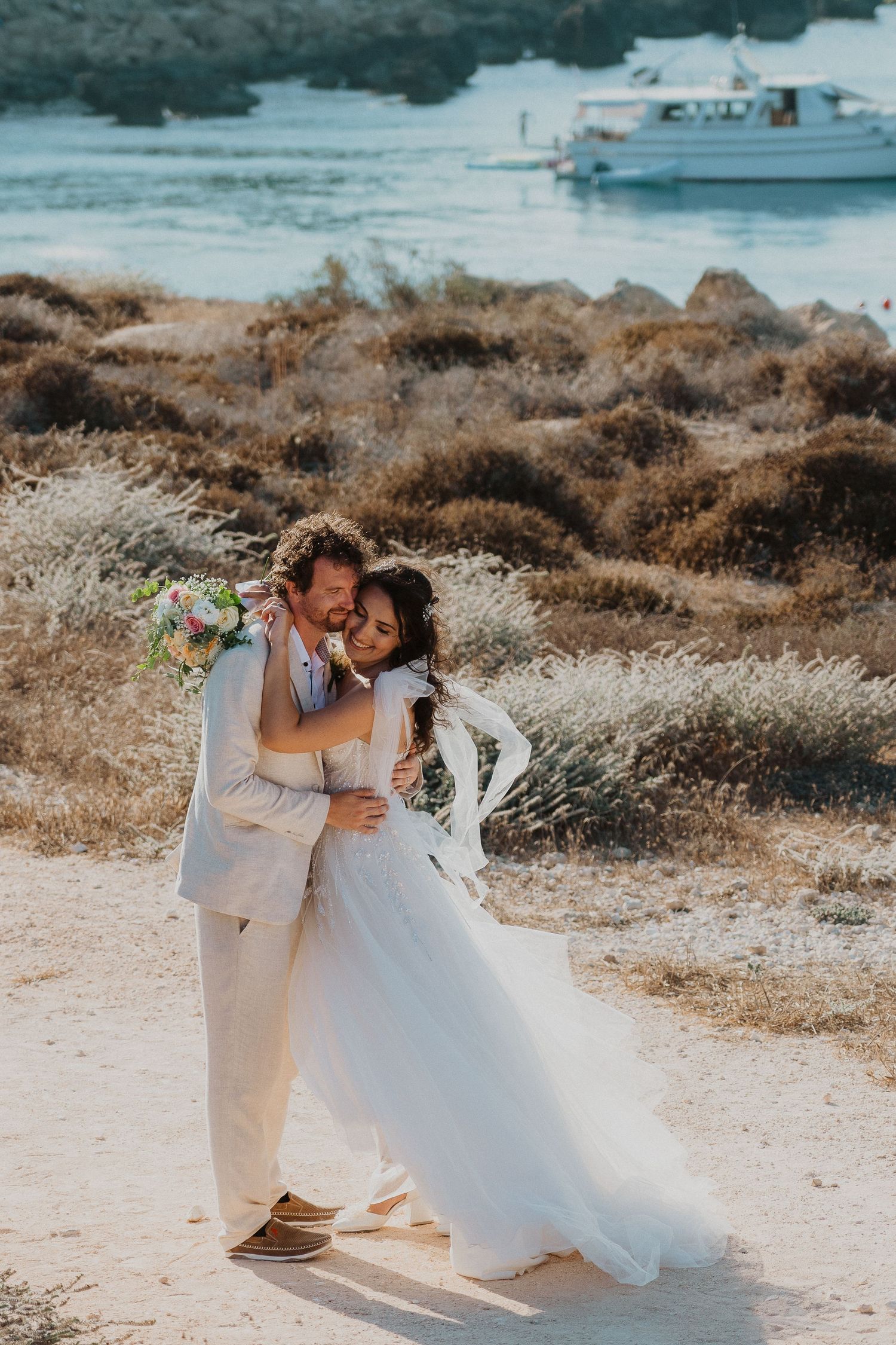 A loving couple shares a romantic moment together in wedding attire against a coastal backdrop with a boat in the distance.