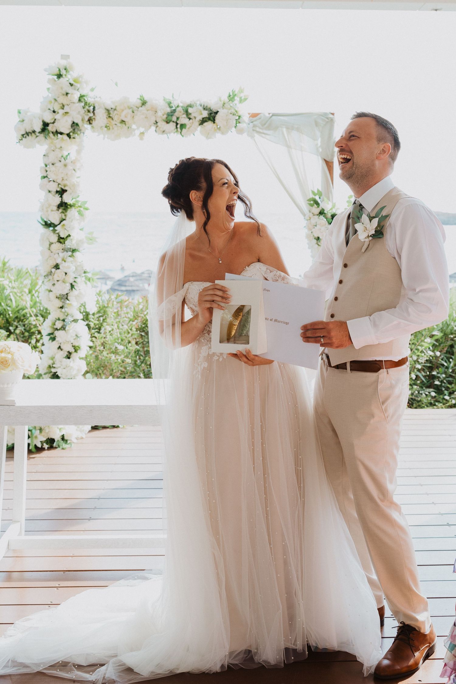 A joyful couple laughs together at an outdoor wedding ceremony beneath a floral arch decorated with white blossoms.