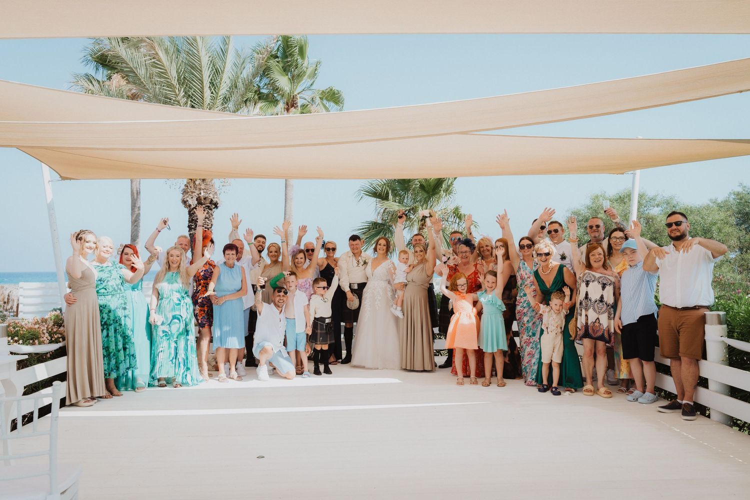 Large group wedding photo of guests celebrating under canopy with ocean backdrop.