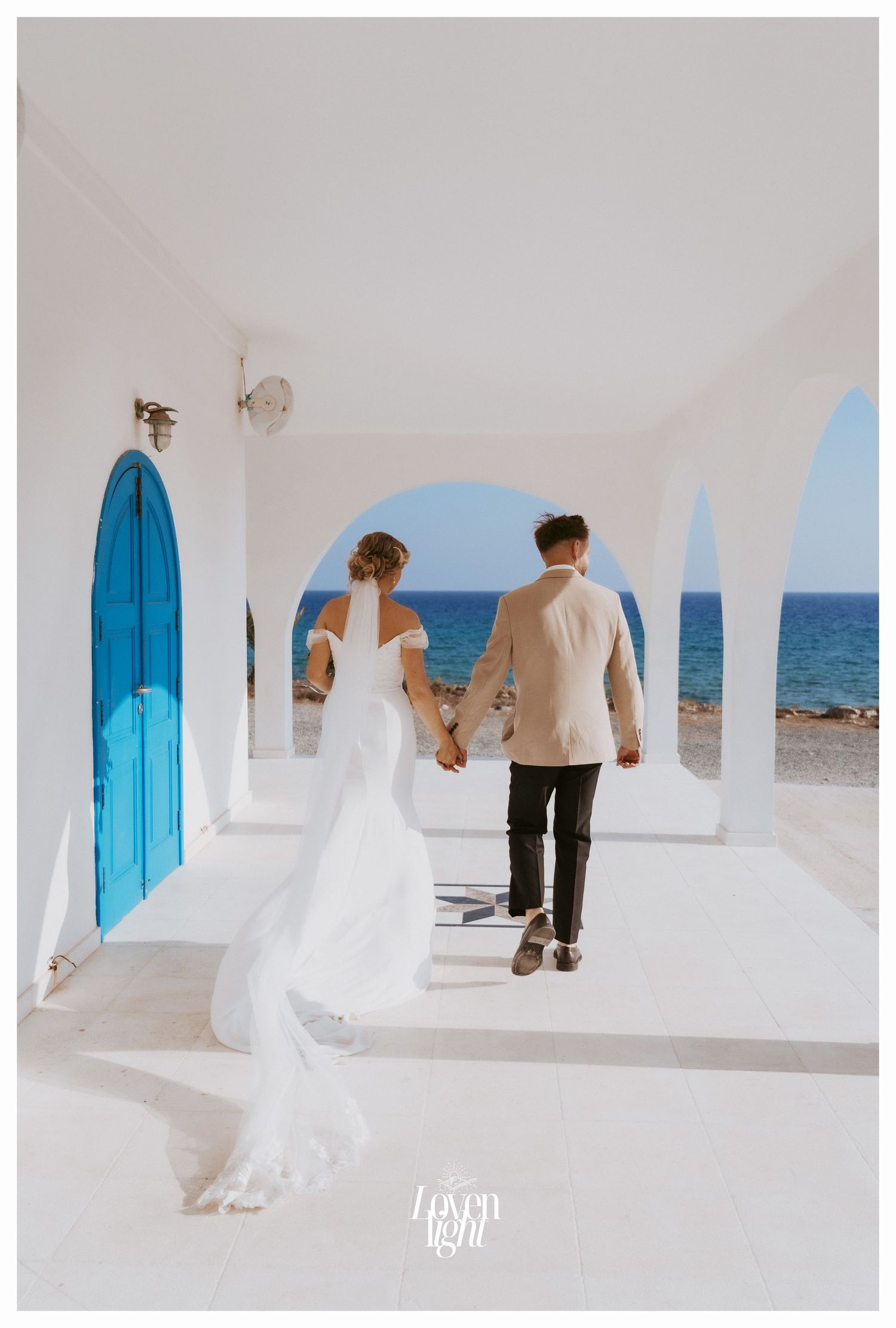 A scenic wedding photo showing a couple walking through white arches with a bright blue door and ocean view in Greece.