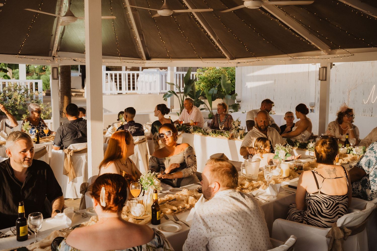 Wedding guests enjoying dinner and conversation at round tables under covered outdoor venue.