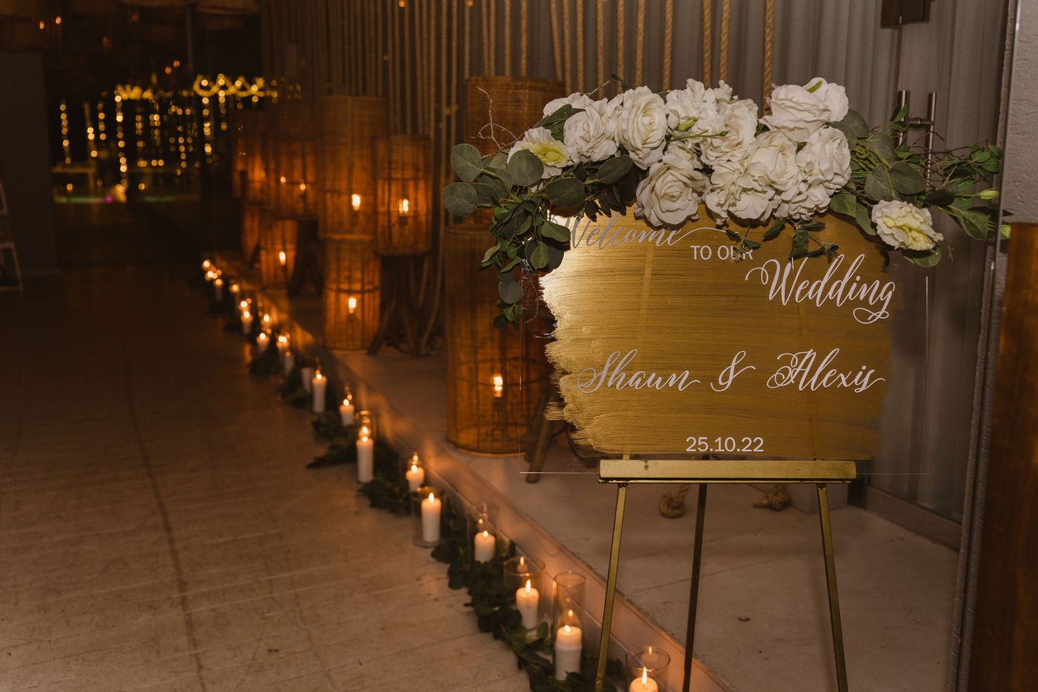 Wedding welcome sign with white floral arrangement illuminated by candlelit pathway.
