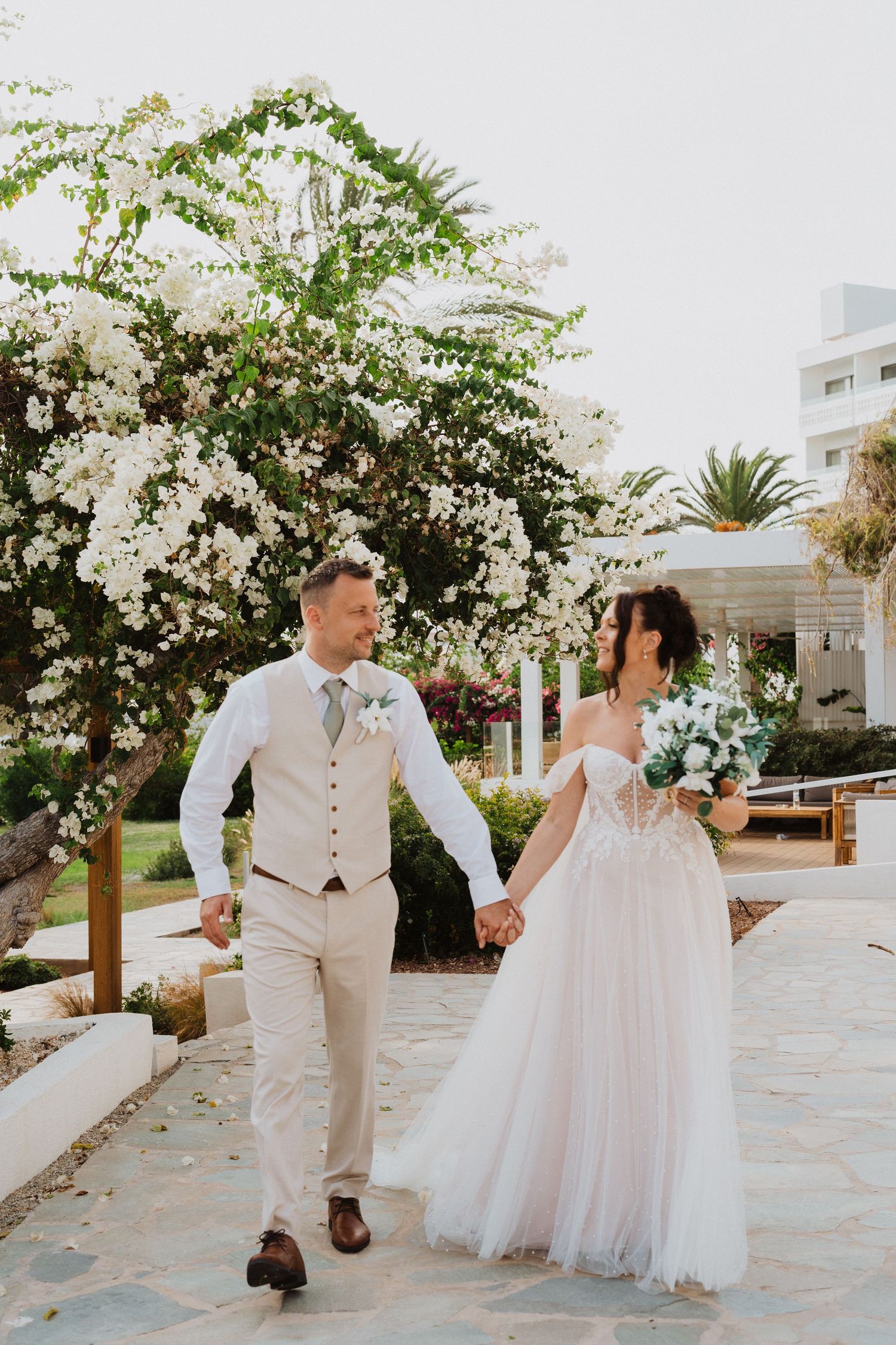 Newlywed couple walks hand in hand through a garden path with blooming white bougainvillea flowers and palm trees.