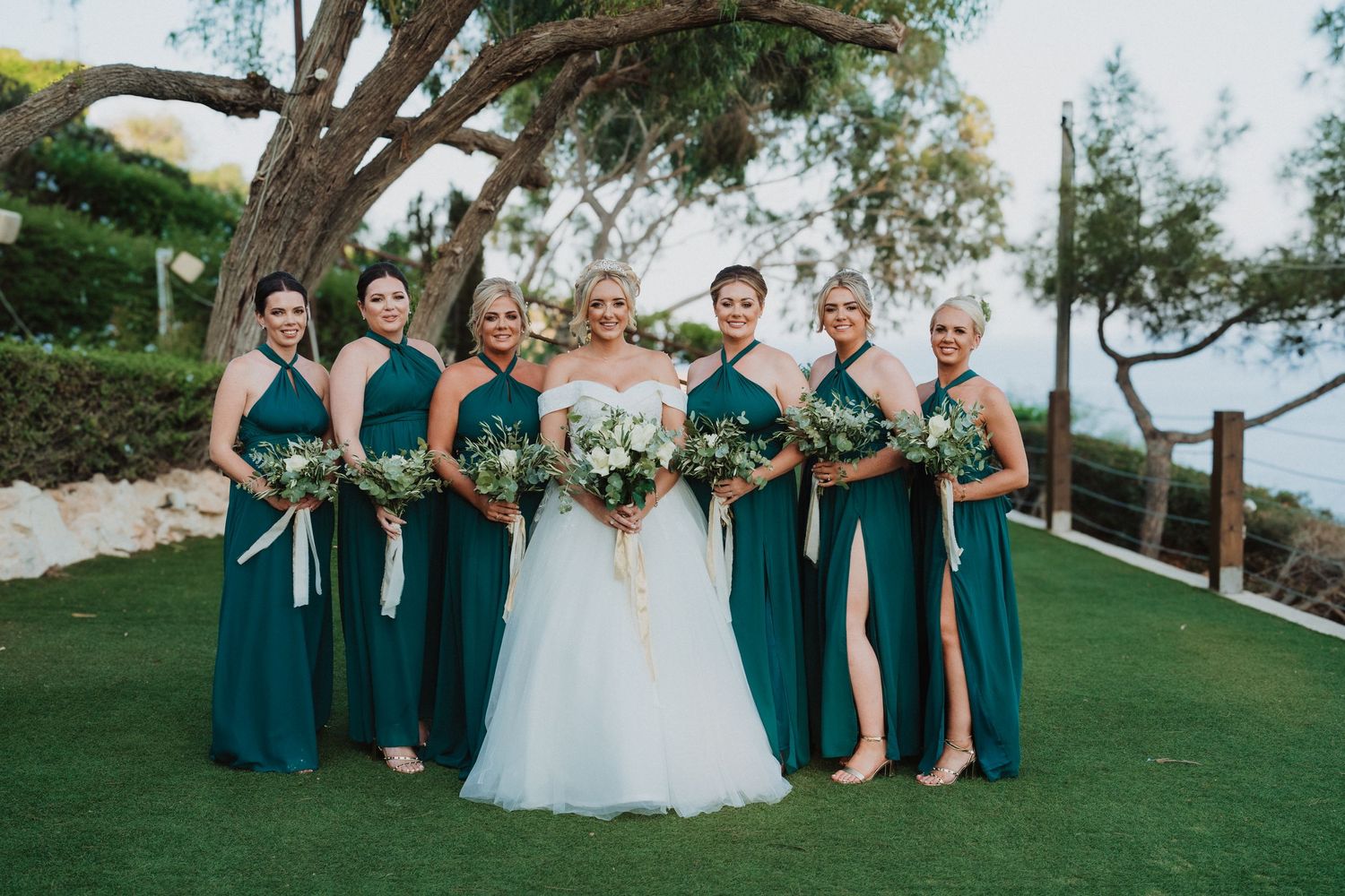 Wedding party in emerald green dresses posing on a manicured lawn with ocean views.