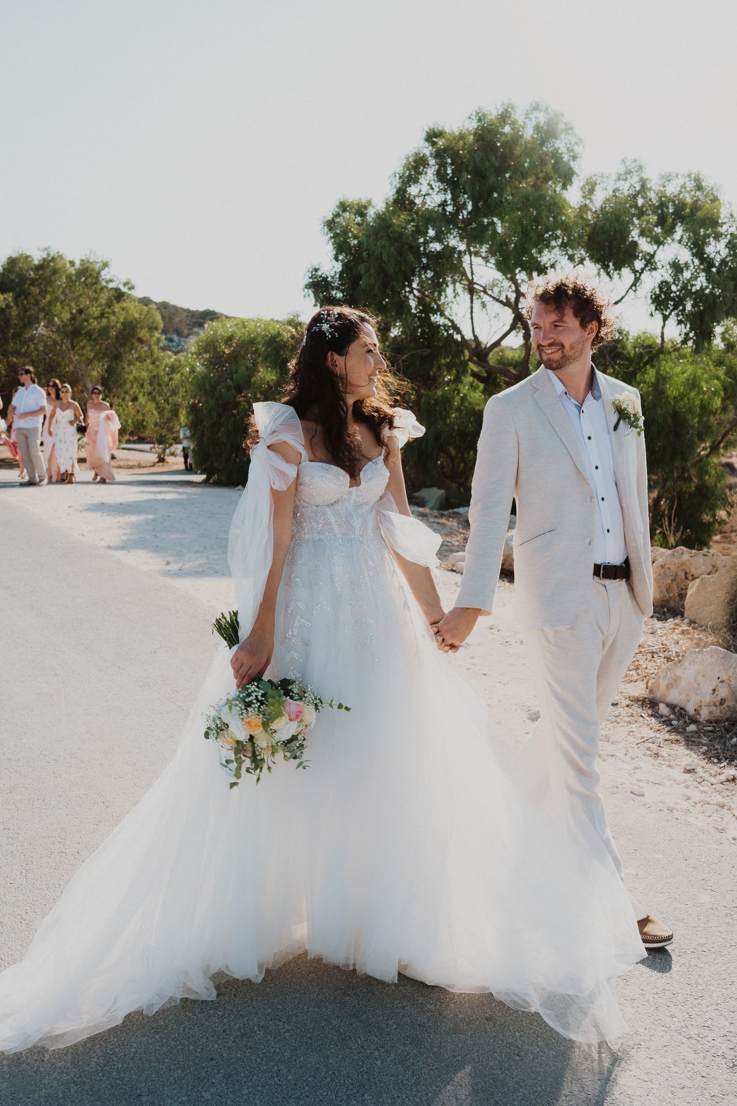 Newlyweds walking together on a path, bride in flowing white dress holding a bouquet.
