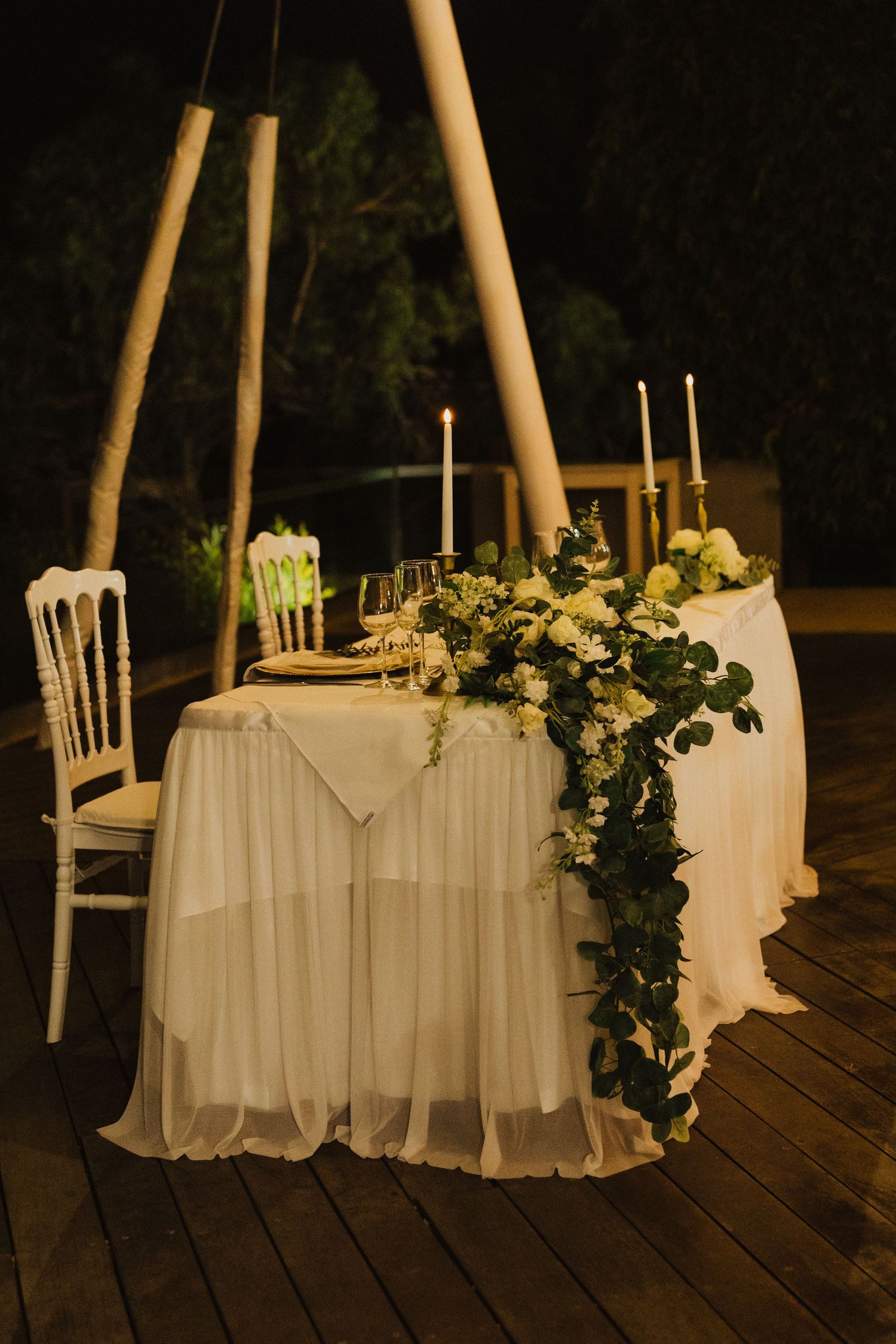 Elegant wedding table setting with white linens and floral garland on wooden deck under pole supports at night.
