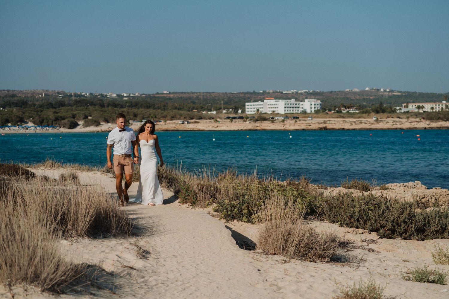 A newlywed couple walks along a coastal path with turquoise waters and buildings in the background.