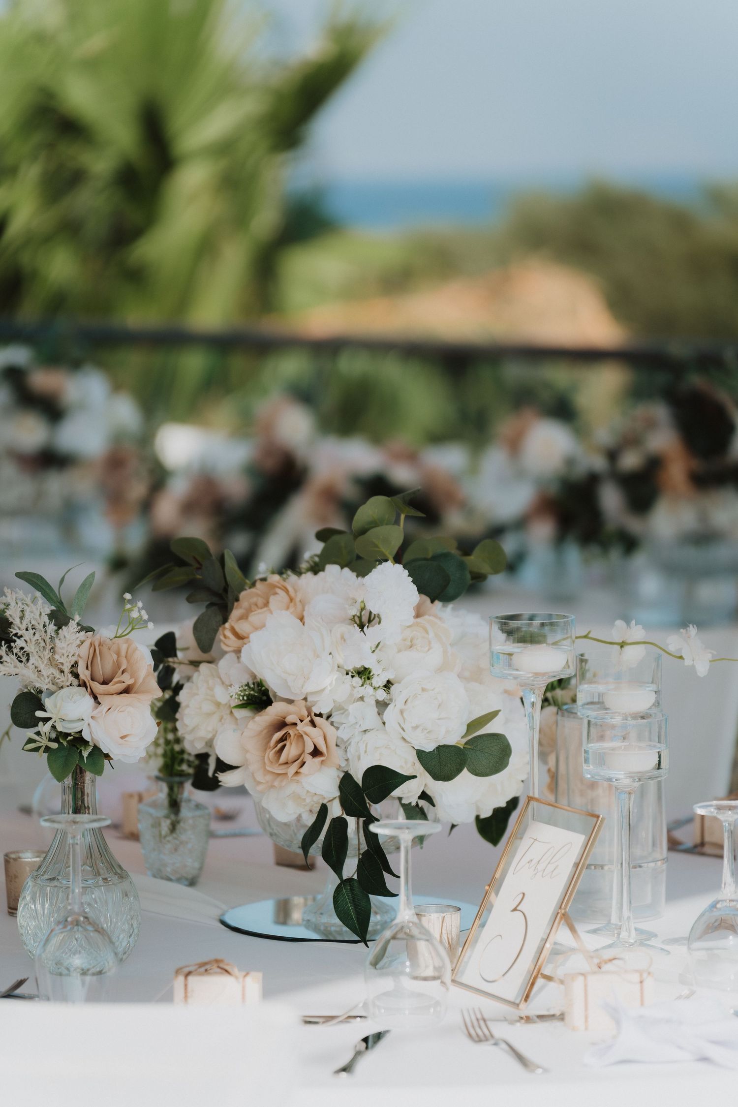 Romantic floral centerpieces with blush roses and eucalyptus on white tablecloths at seaside venue.