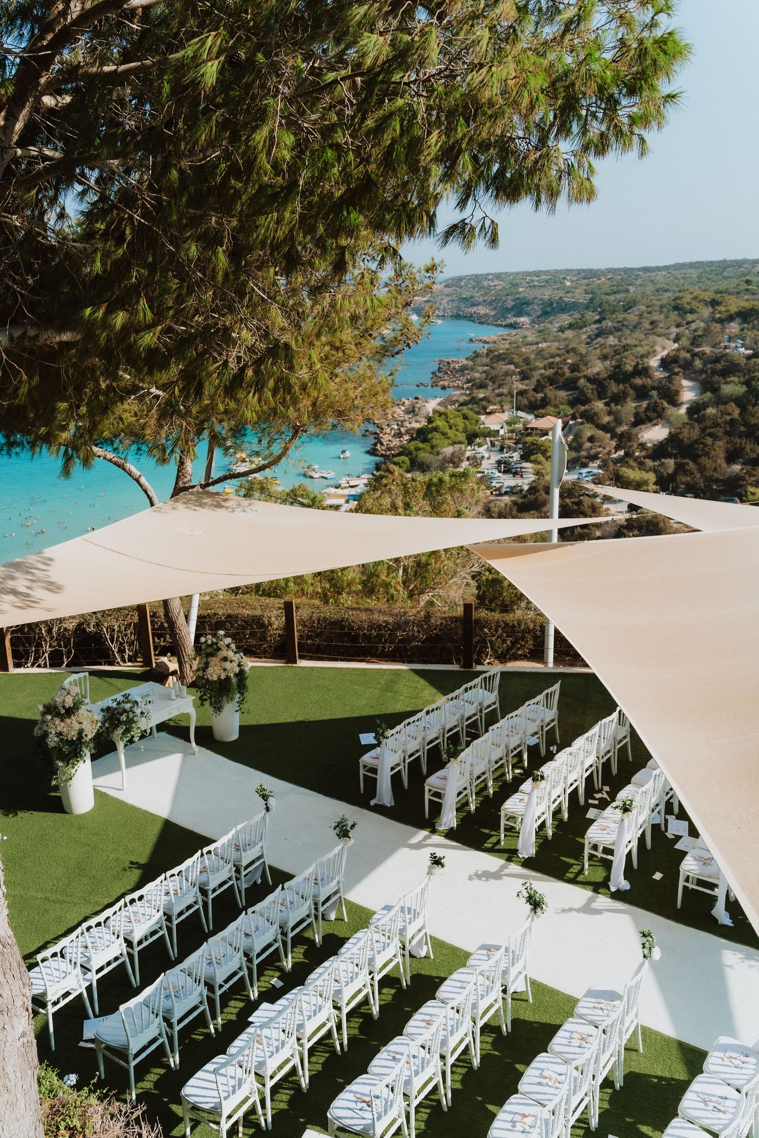 Elegant outdoor wedding venue with white chairs and shade sails set against a coastal Mediterranean backdrop.