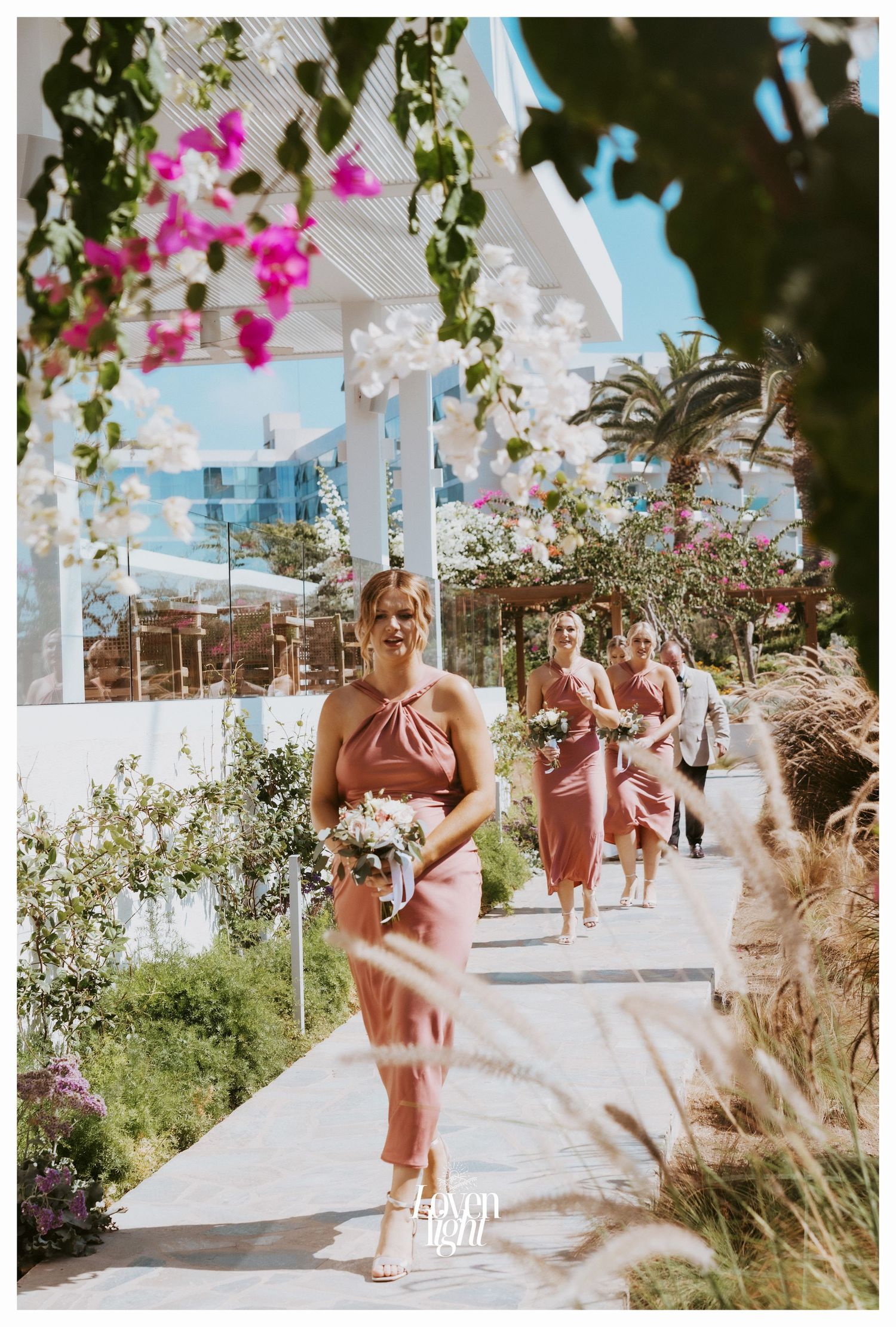 Bridesmaids in coral dresses walk down flower-lined outdoor path under blue sky.