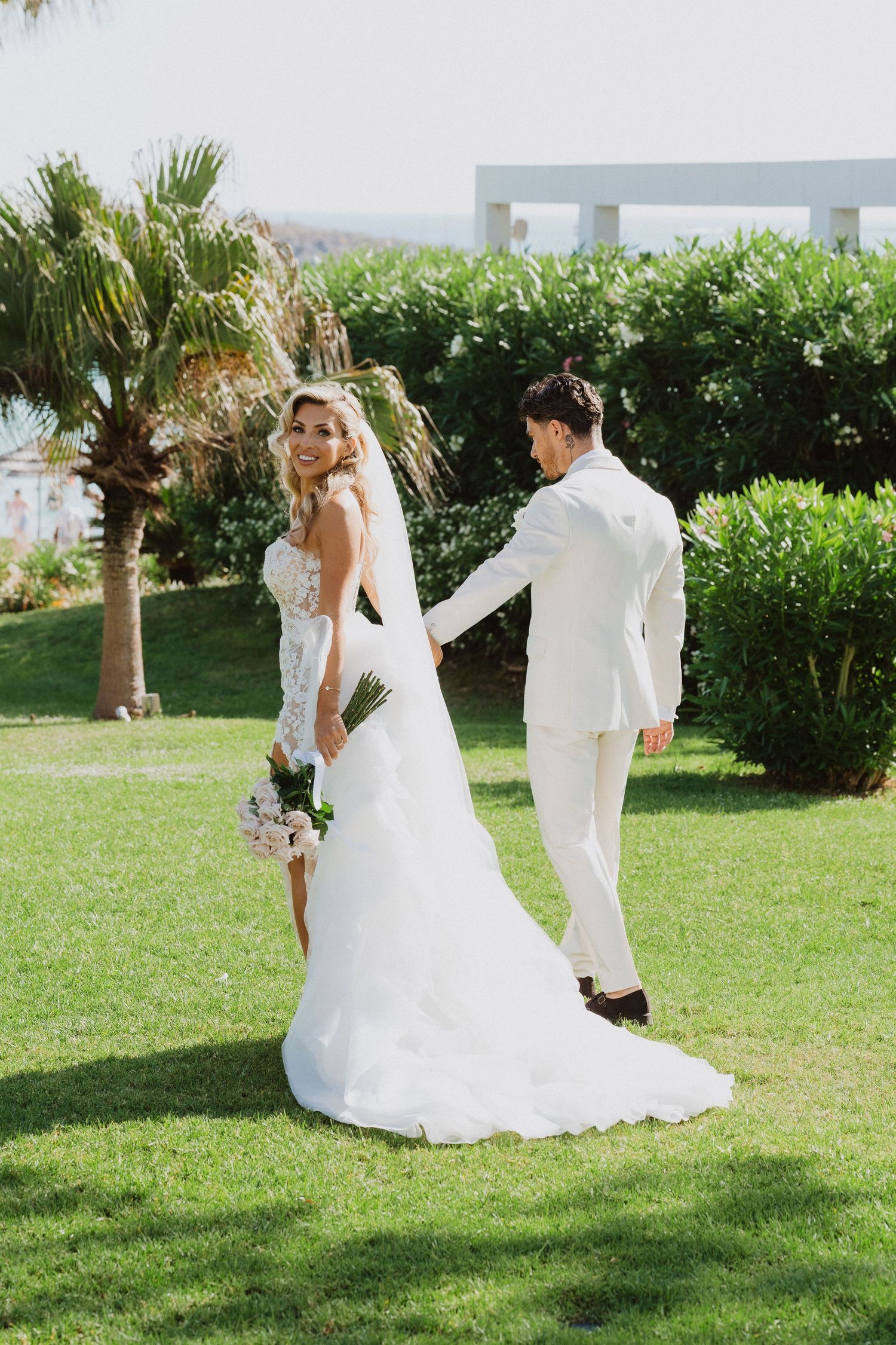 A couple in white wedding attire dance playfully on a lush green lawn with palm trees in the background.