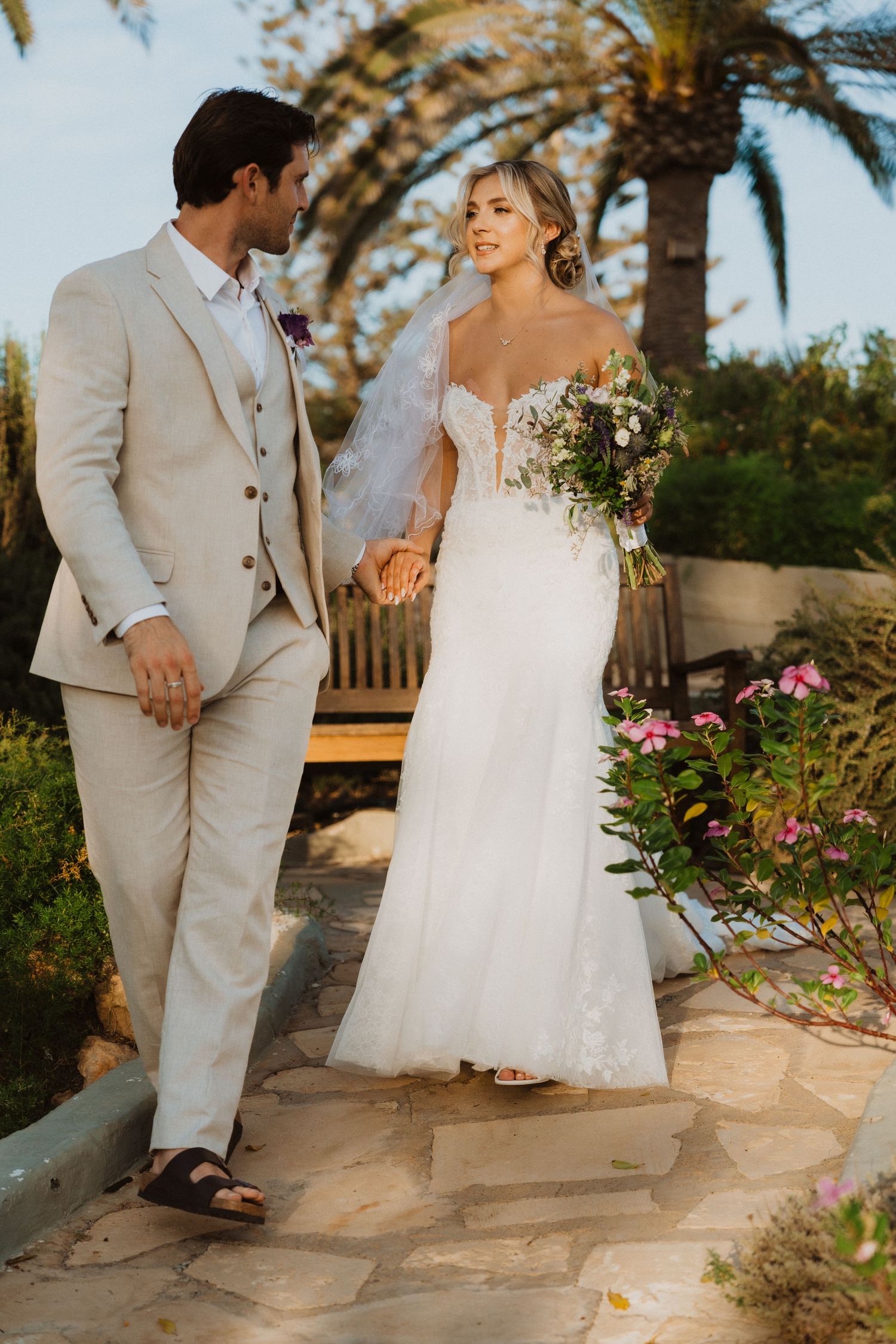 Couple in beachwear and wedding attire walking along a stone path with tropical plants.