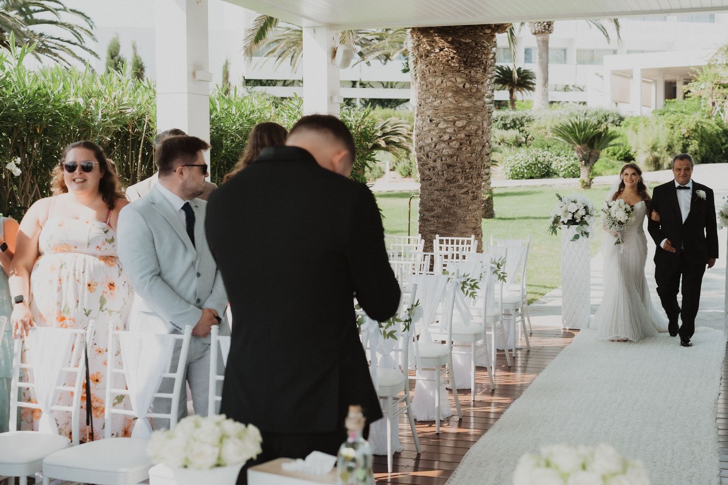 Bride walking down aisle escorted by father toward groom at outdoor wedding ceremony venue.