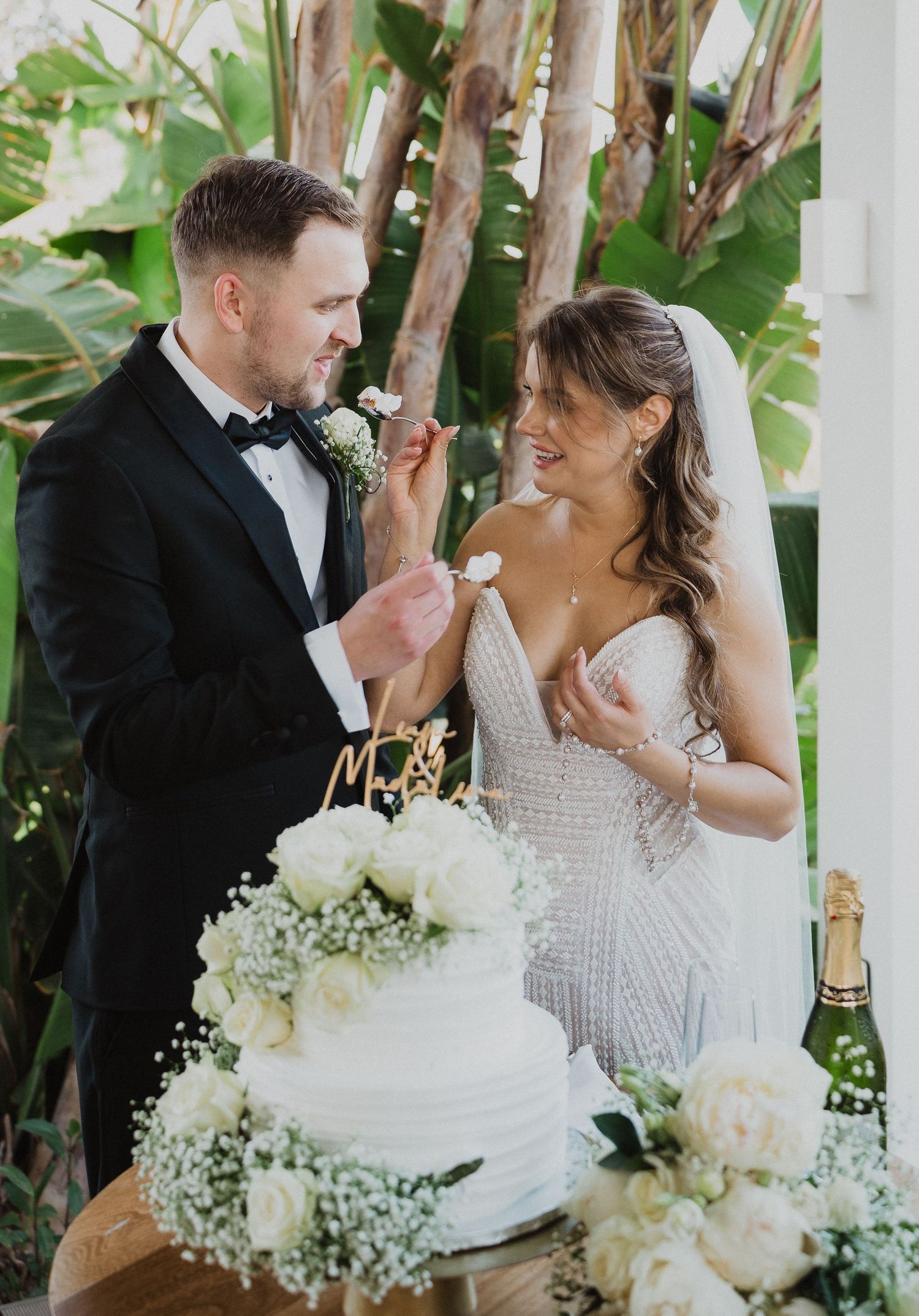 Bride and groom cutting white floral wedding cake, groom in tuxedo feeding bride in beaded gown outdoors.