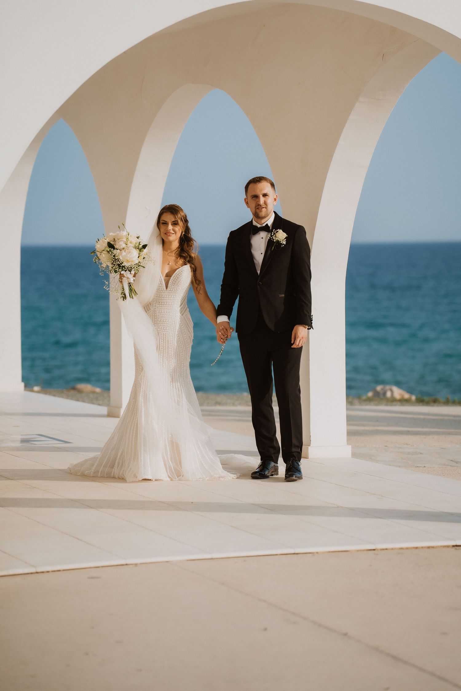 Bride and groom posing under white arches with bouquet, overlooking a stunning blue sea.