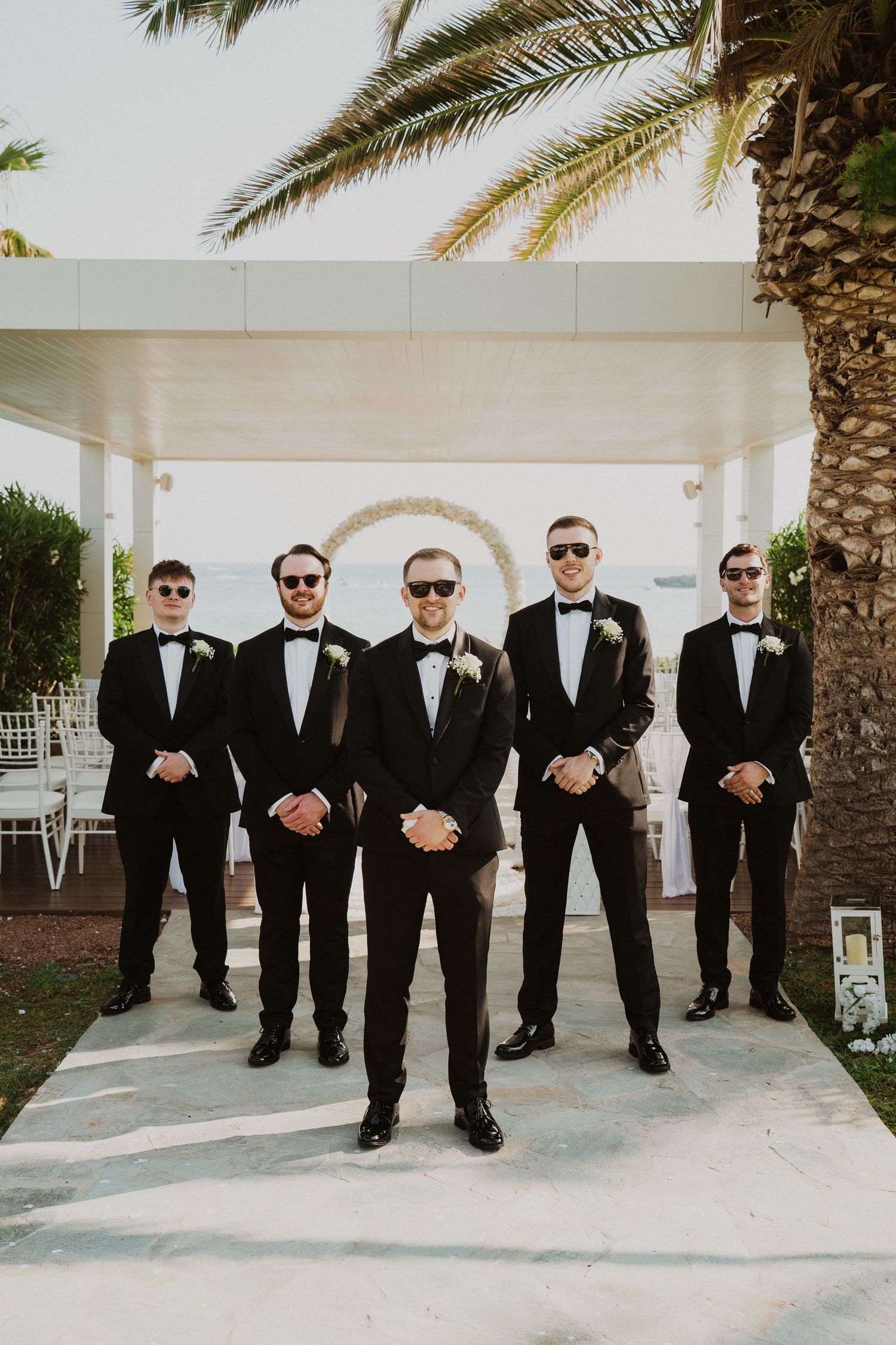Five groomsmen in matching black tuxedos and sunglasses pose confidently outdoors at a seaside wedding venue.