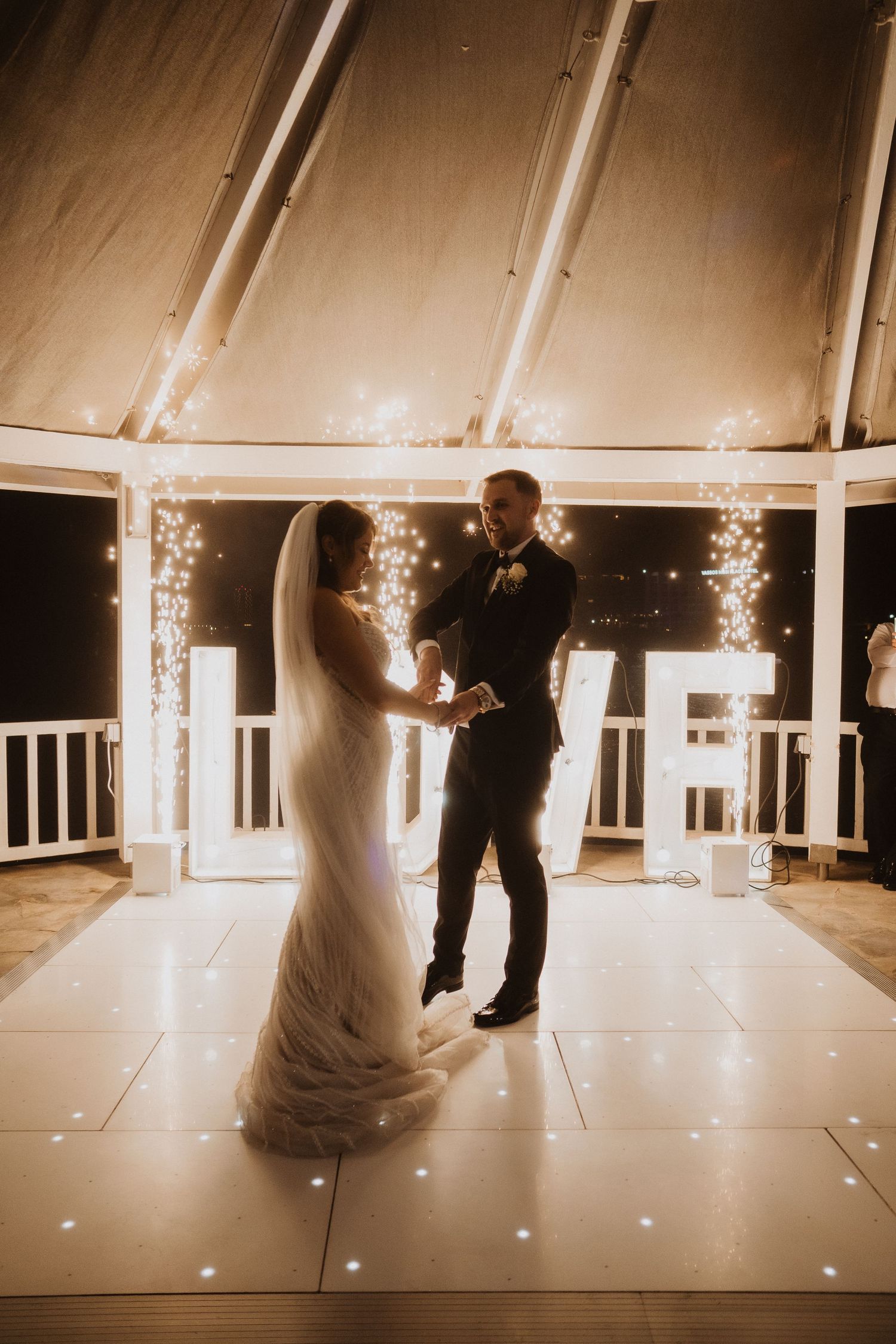 Bride and groom sharing first dance on illuminated white floor with LOVE letters and fairy lights backdrop.