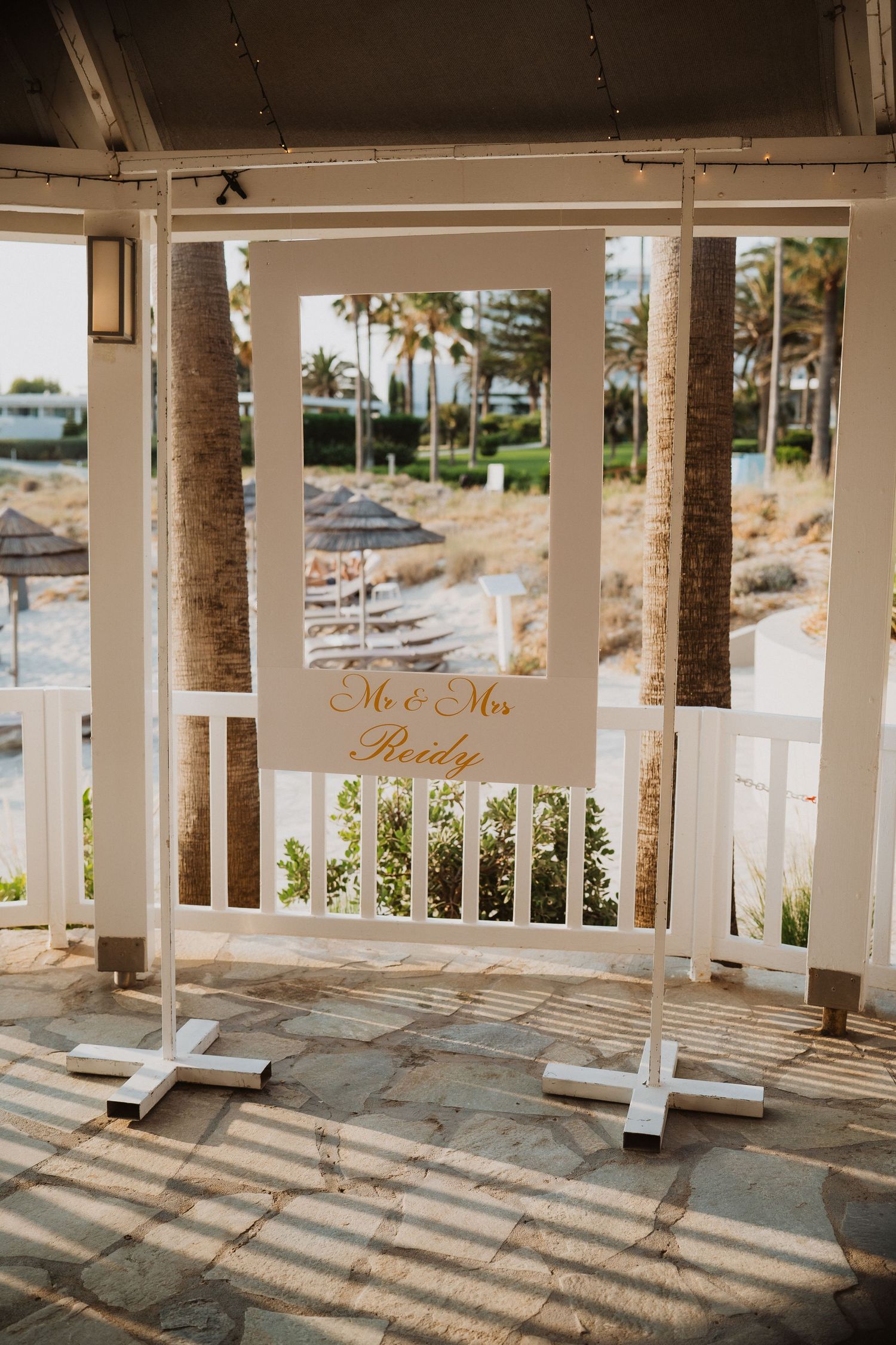 White mirror wedding welcome sign reading Mr and Mrs Brady displayed at outdoor beachside wedding venue with palm trees.