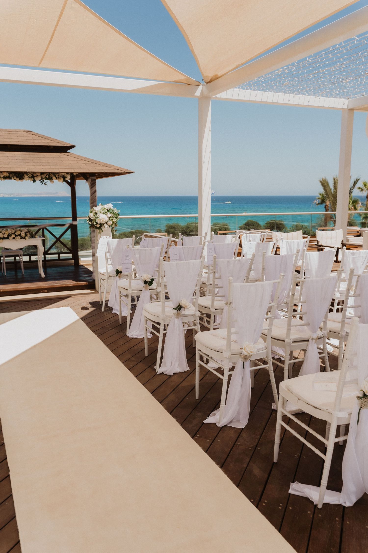 Elegant beachfront wedding reception setup with white chairs and tables under cream umbrellas overlooking turquoise ocean waters.