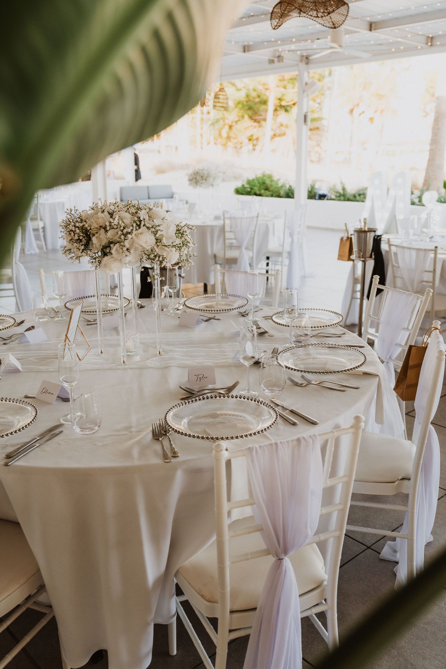 Elegant white wedding reception tables with floral centerpieces and beaded charger plates in an outdoor setting.