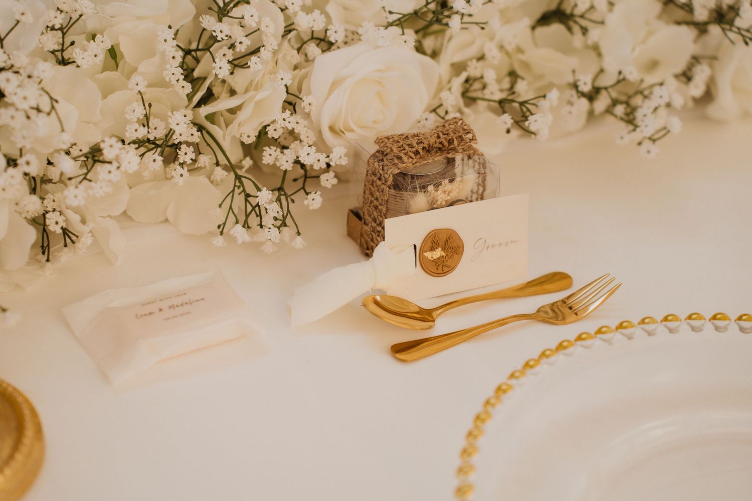 White floral centerpiece with gold cutlery, wax seal card, and pearl-beaded charger plate setting.