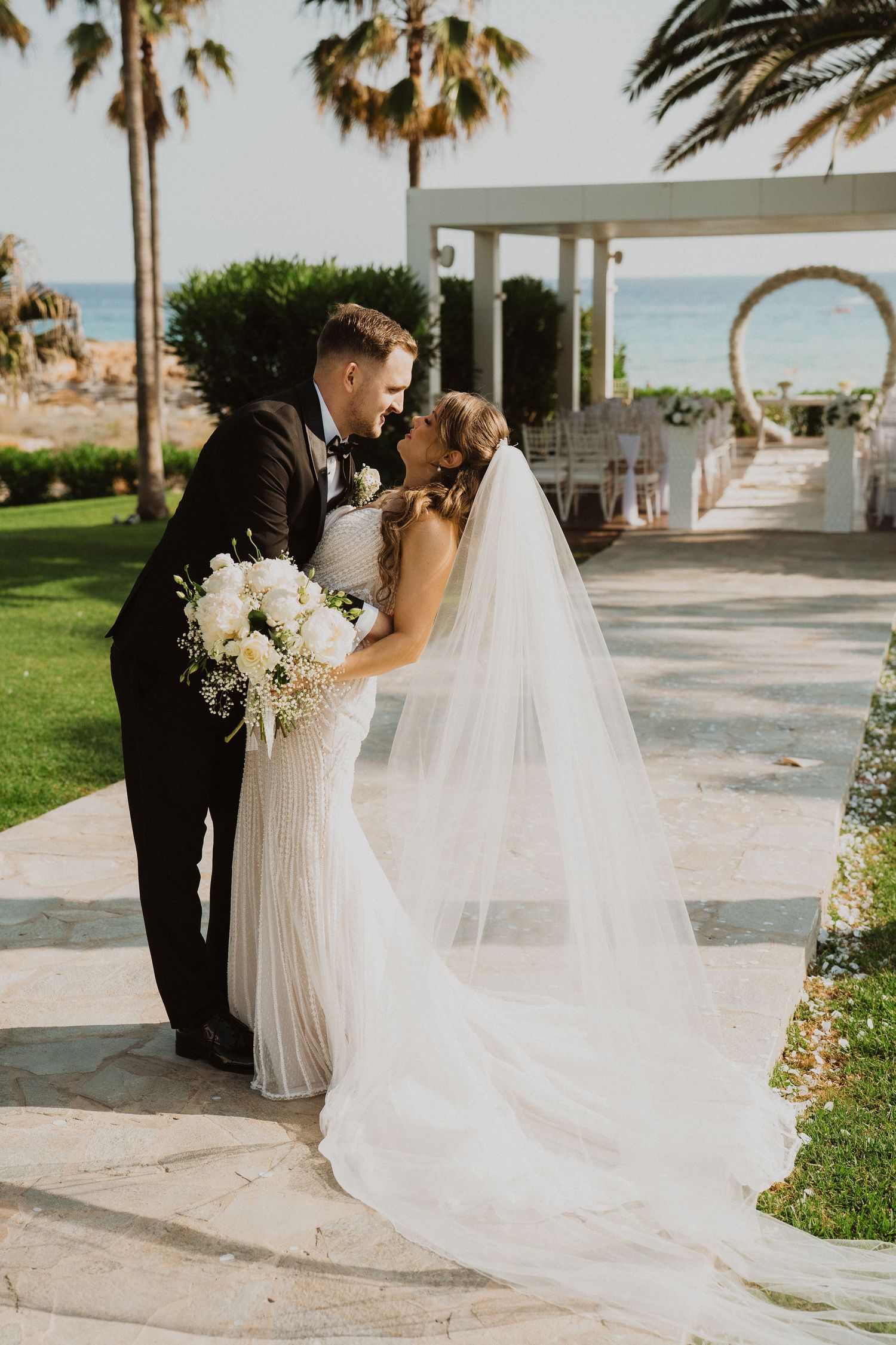 Bride and groom sharing a kiss on a stone path with palm trees and ocean backdrop at a beachside wedding venue.