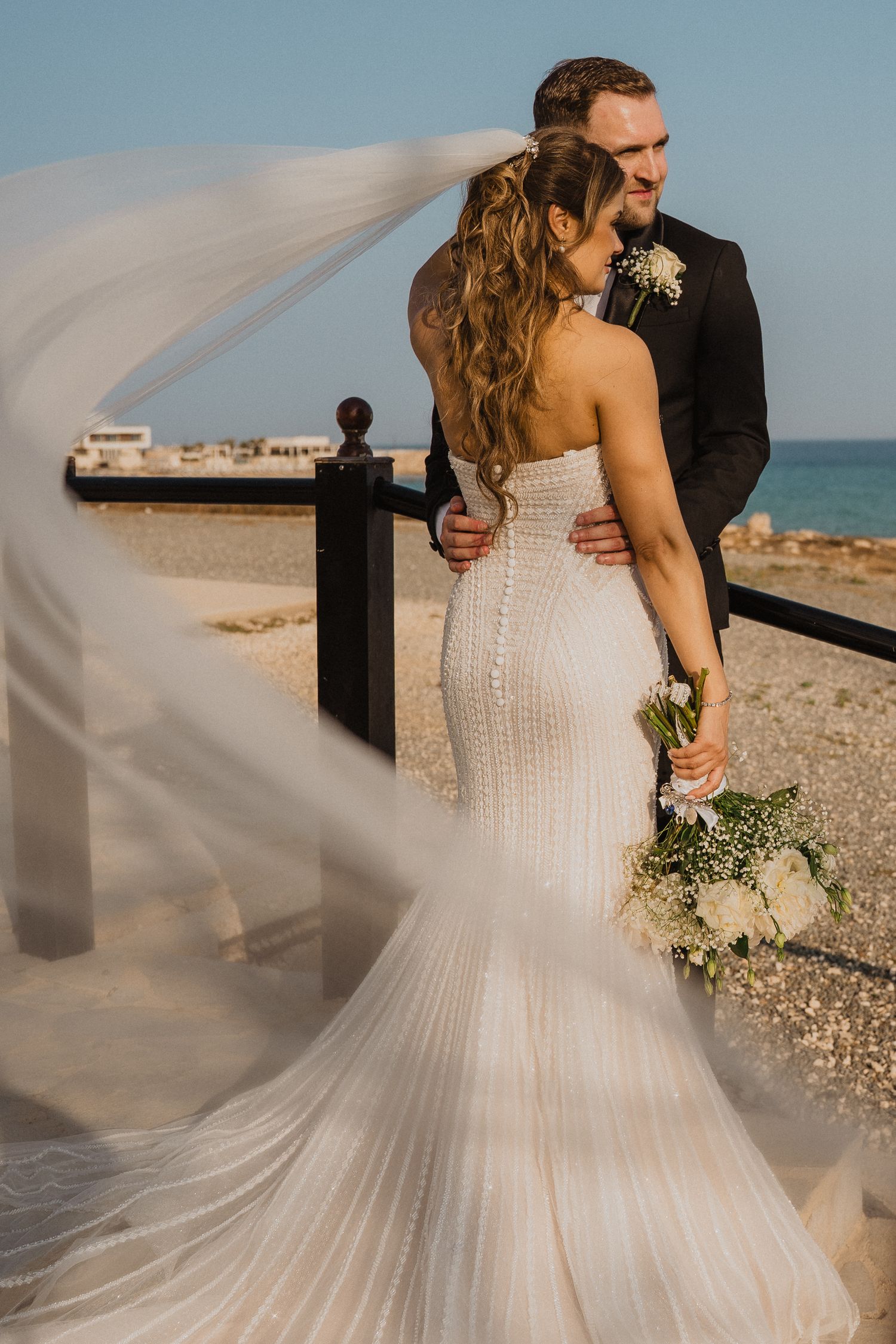 Bride in beaded gown holds white bouquet as veil billows in wind near ocean with groom embracing her.