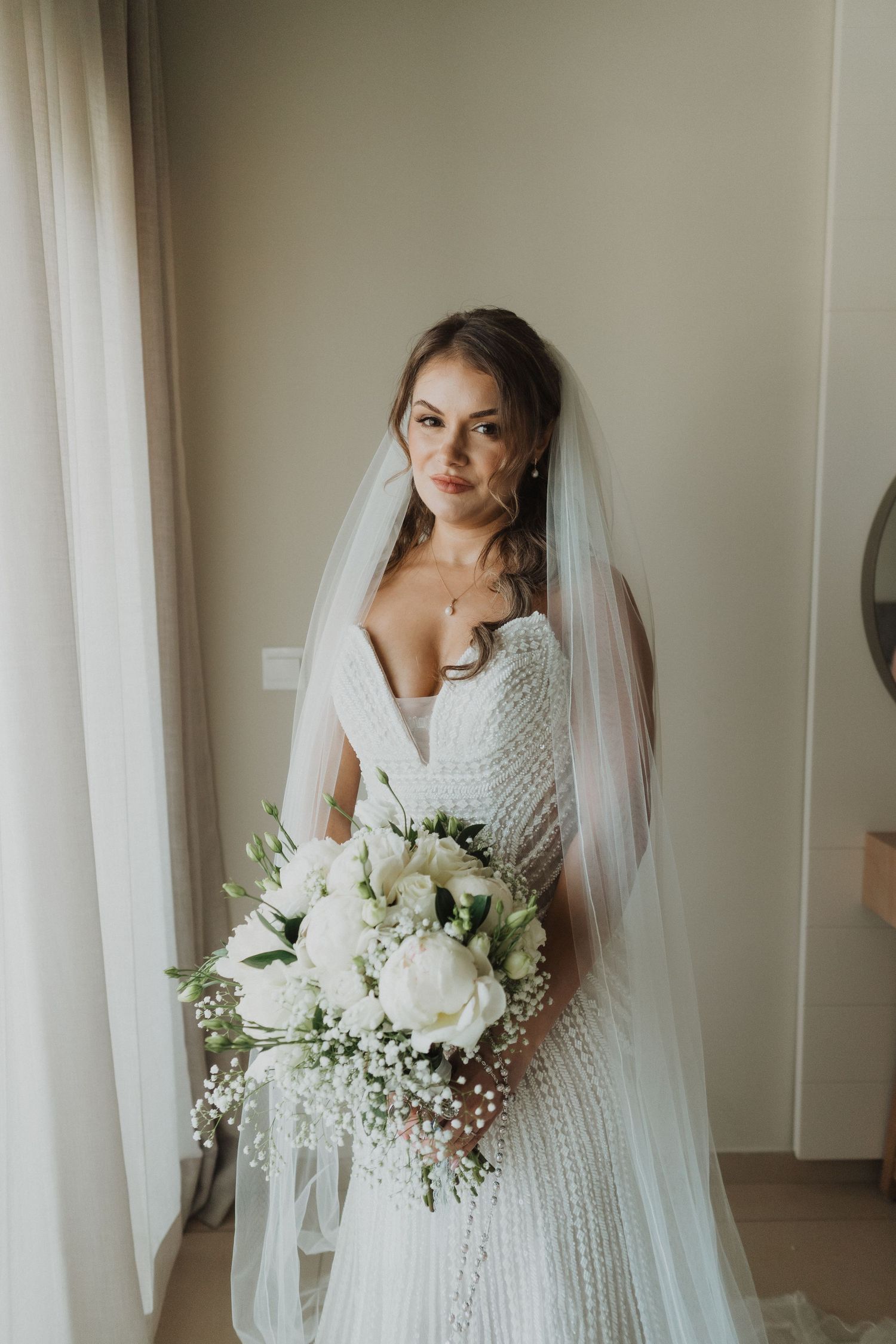 Elegant bride in beaded wedding dress holding white rose bouquet with flowing veil indoors.