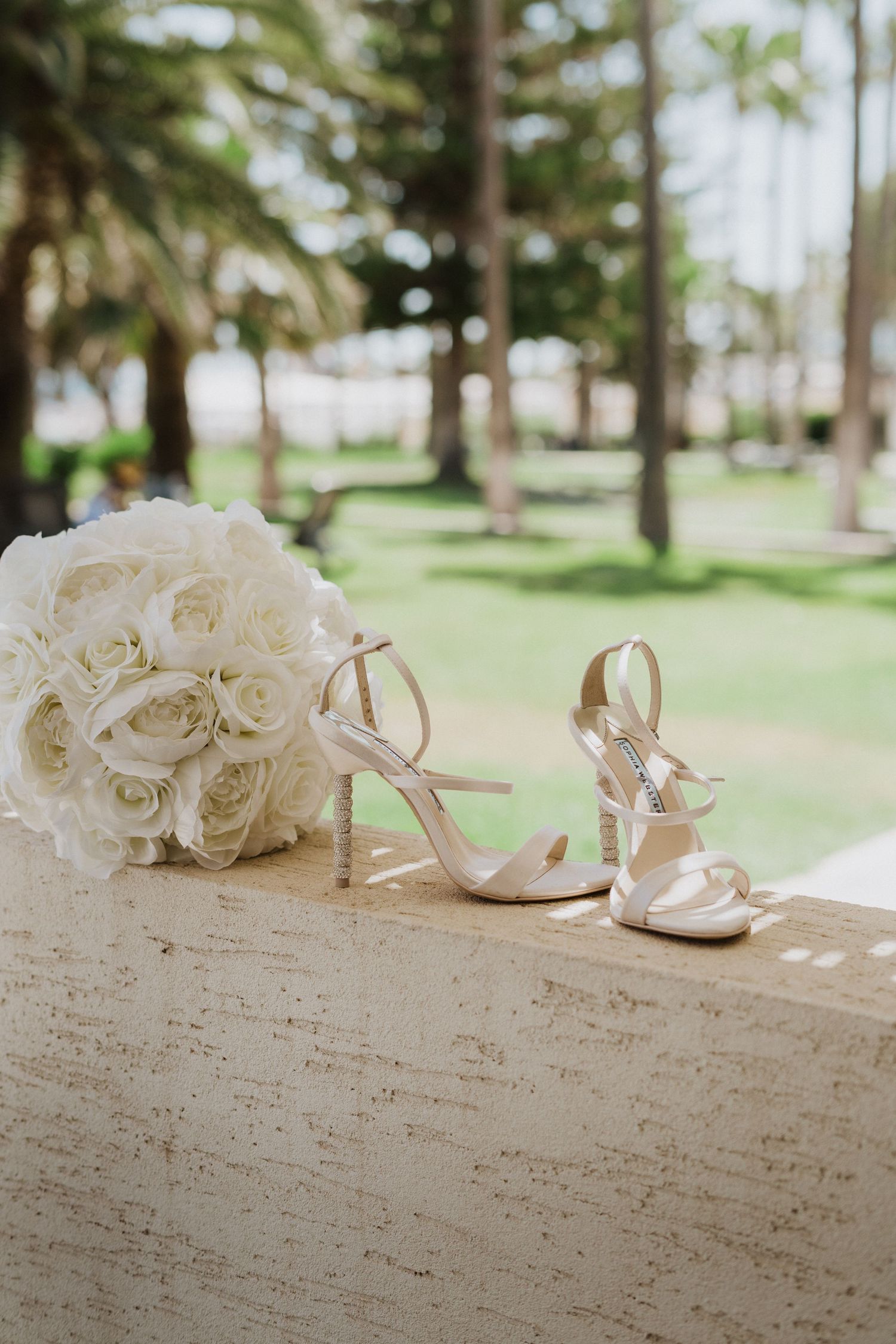 White bridal bouquet and elegant silver wedding shoes arranged on a textured surface with palm trees in background.