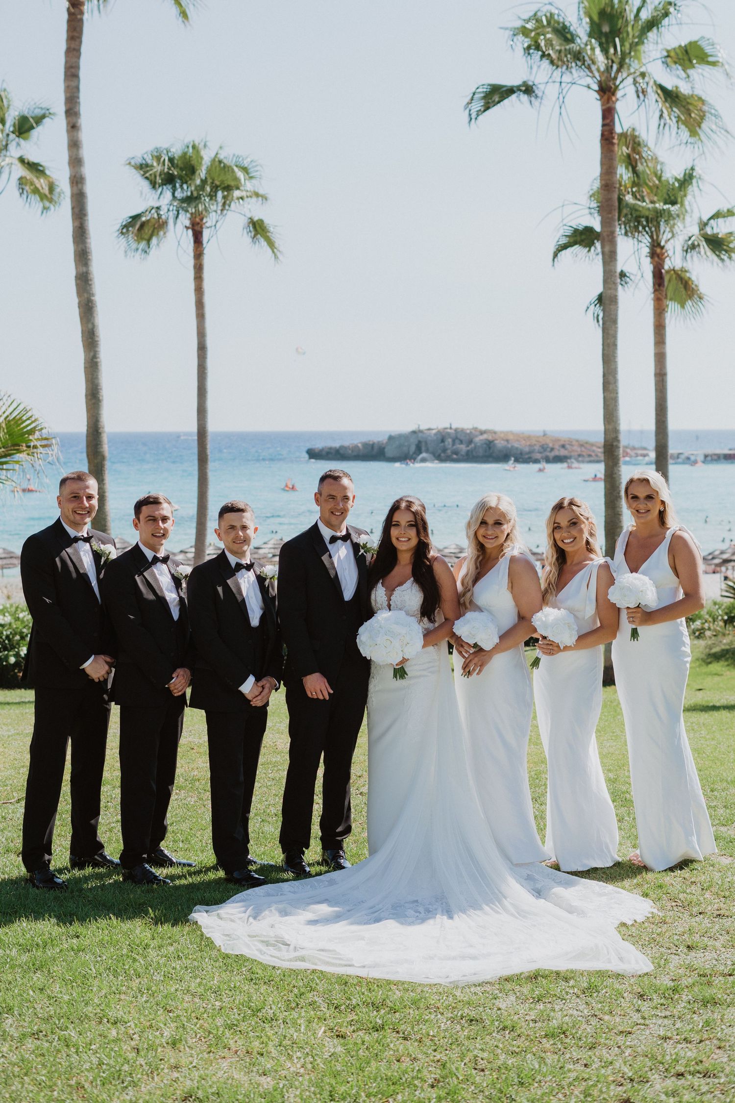 Wedding party in black and white attire posed on grass with ocean and palm trees in background.